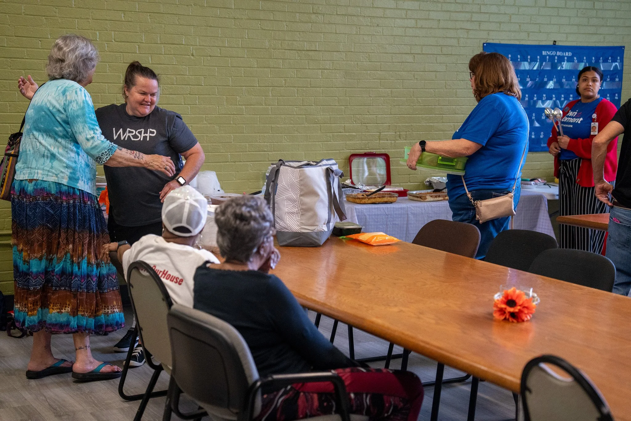 People gathered in a room around a table with food and utensils, with a green brick wall in the background. Some are sitting, and others are standing and talking.