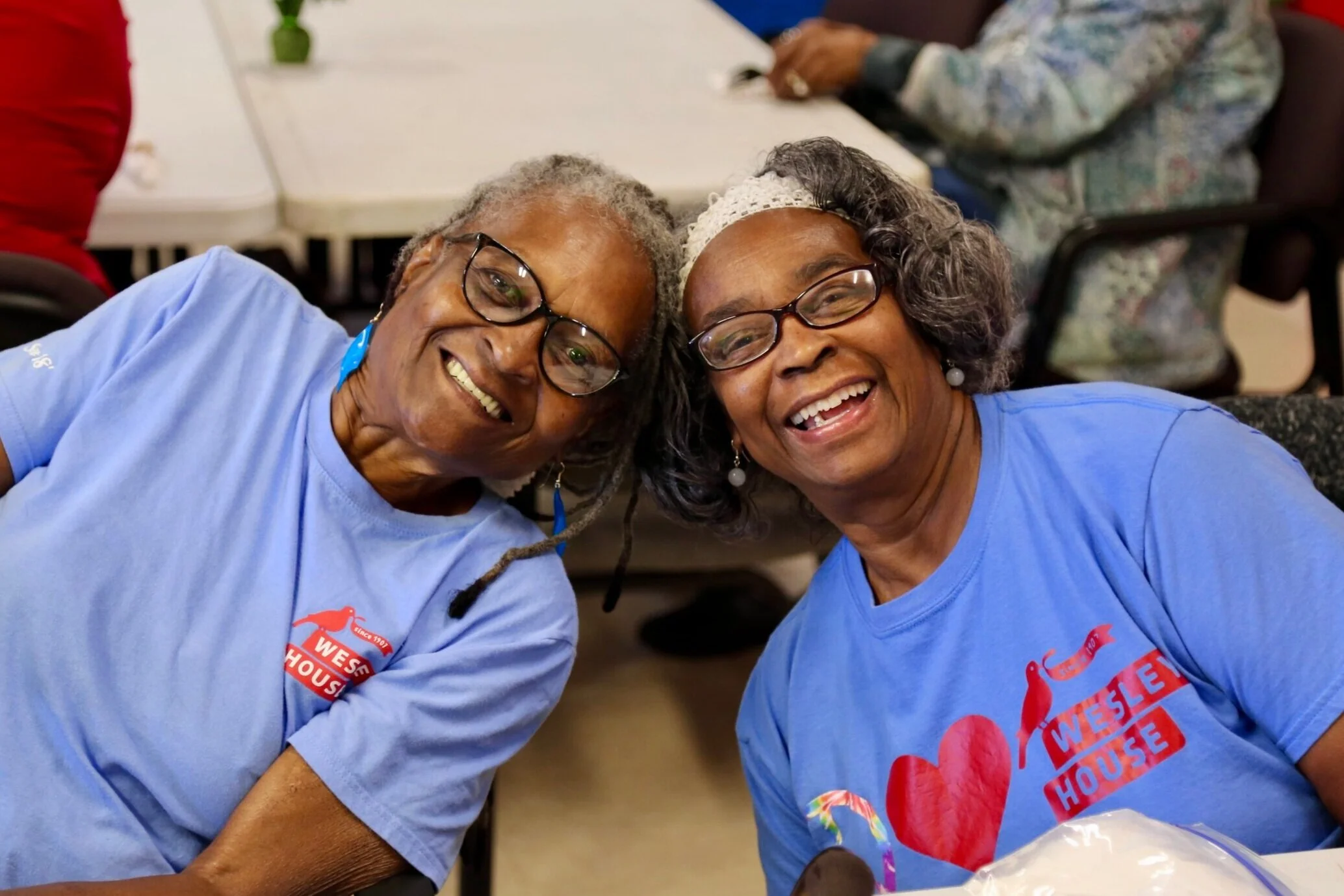 Two smiling older women with glasses, wearing blue T-shirts with 'Weston House' logos, sitting close together at a table in a social setting.