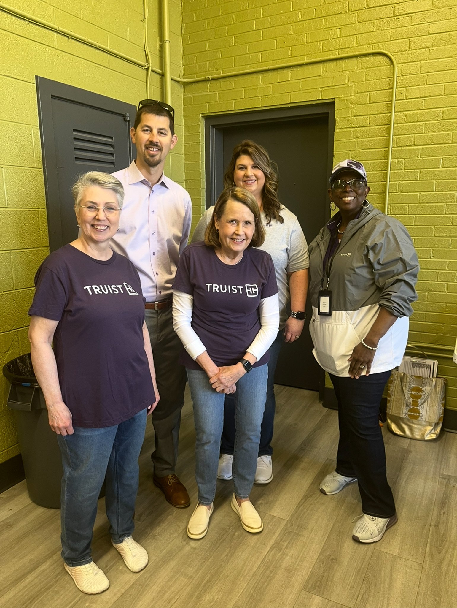 Group of five diverse people standing together indoors, smiling, with two women wearing purple 'TRUIST' t-shirts.