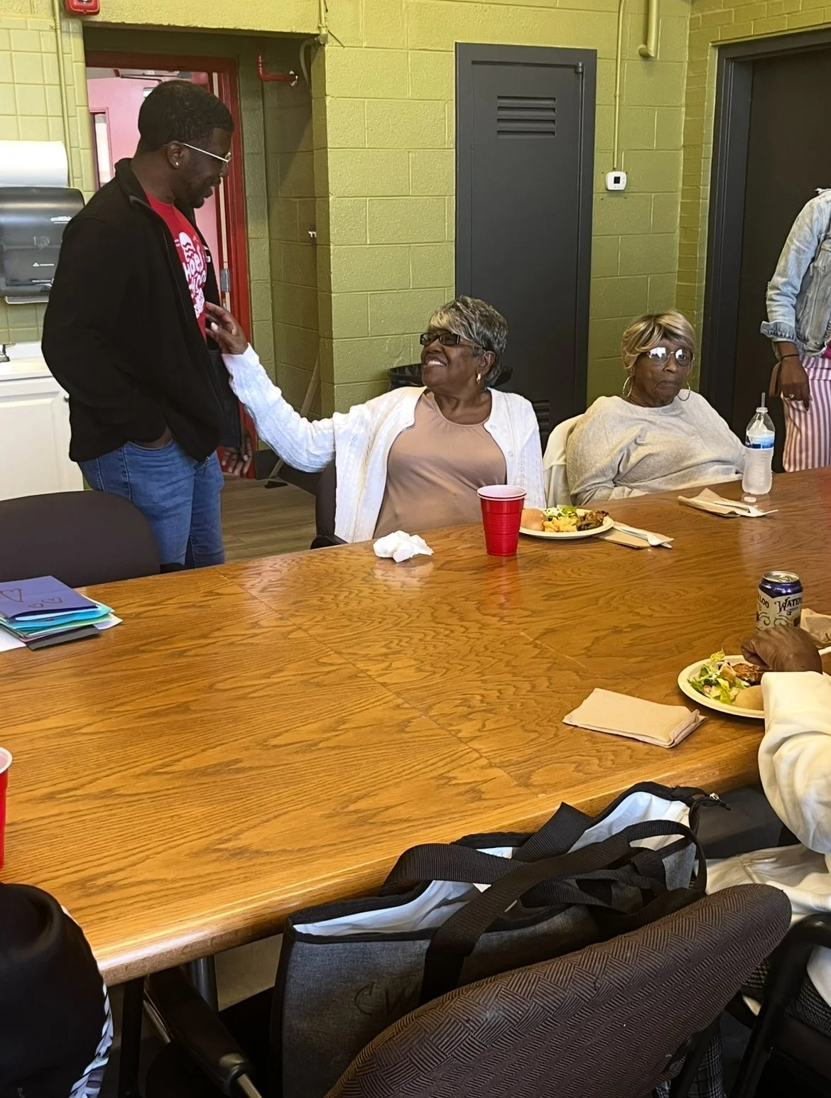 A woman in a white sweater touching the chest of a man in a black jacket and red shirt, sitting at a lunch table. Other women are seated nearby, with plates of food, drinks, and a water bottle. The setting appears to be a casual indoor gathering or m
