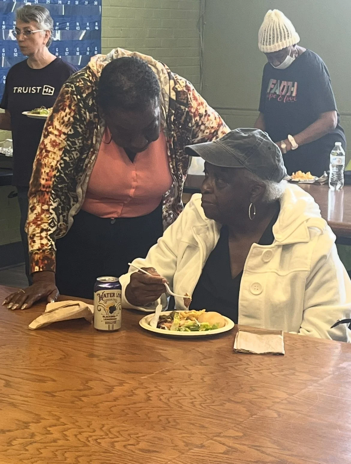 Older woman sitting at a table with a paper plate of food, holding a fork, wearing a white jacket and a cap. Another woman leaning over the table, engaging with her. In the background, two women, one with gray hair and glasses wearing a black T-shirt
