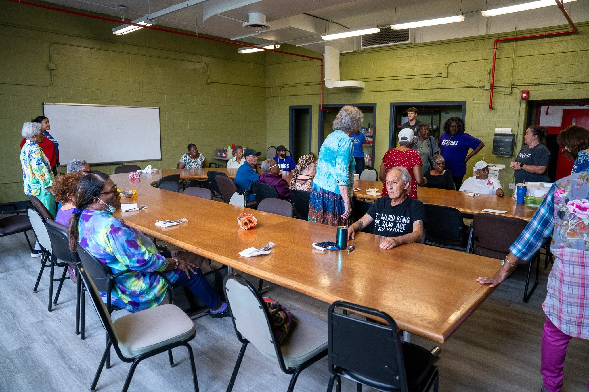 A diverse group of elderly people gathering in a community room with green walls and a wooden table, some sitting and some standing, engaging in conversation.