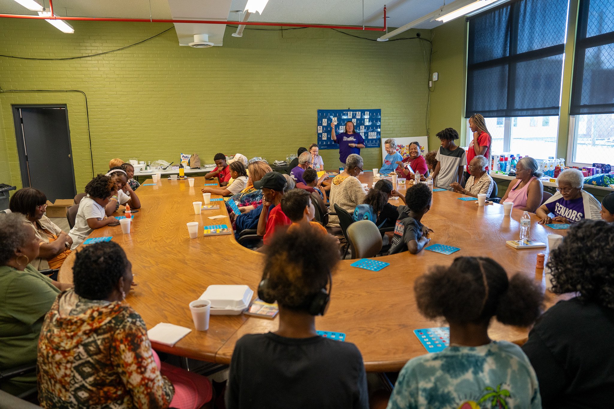 A diverse group of people, including children and elderly, gathered around a large oval table in a community room, participating in a group activity or meeting with some people standing and speaking at the front.
