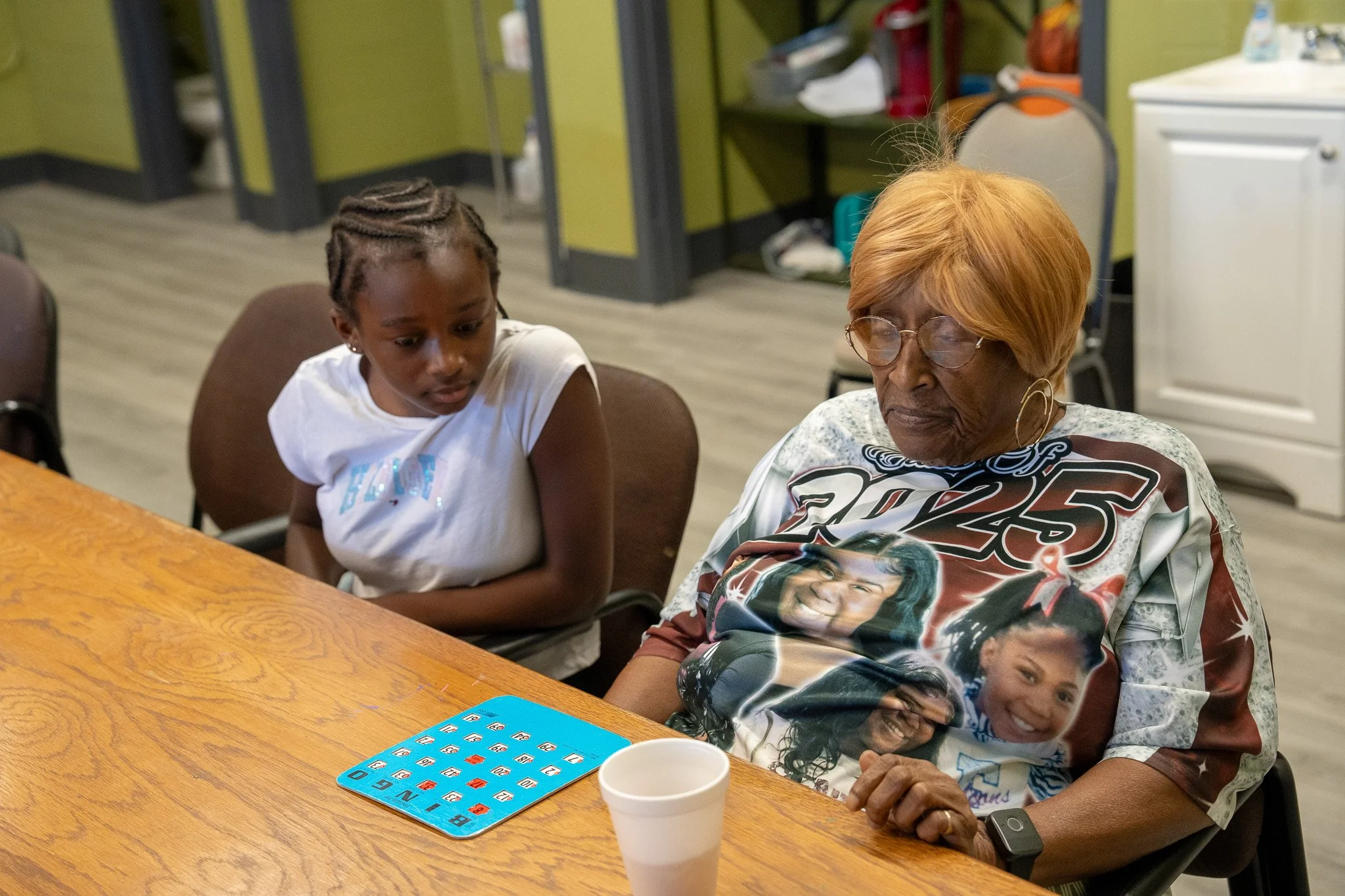A young girl and an elderly woman sitting at a table, looking at a bingo card with brightly colored numbers. The woman is wearing a T-shirt with photographs of three women and the number 2025, while the girl is wearing a white shirt with a shiny desi