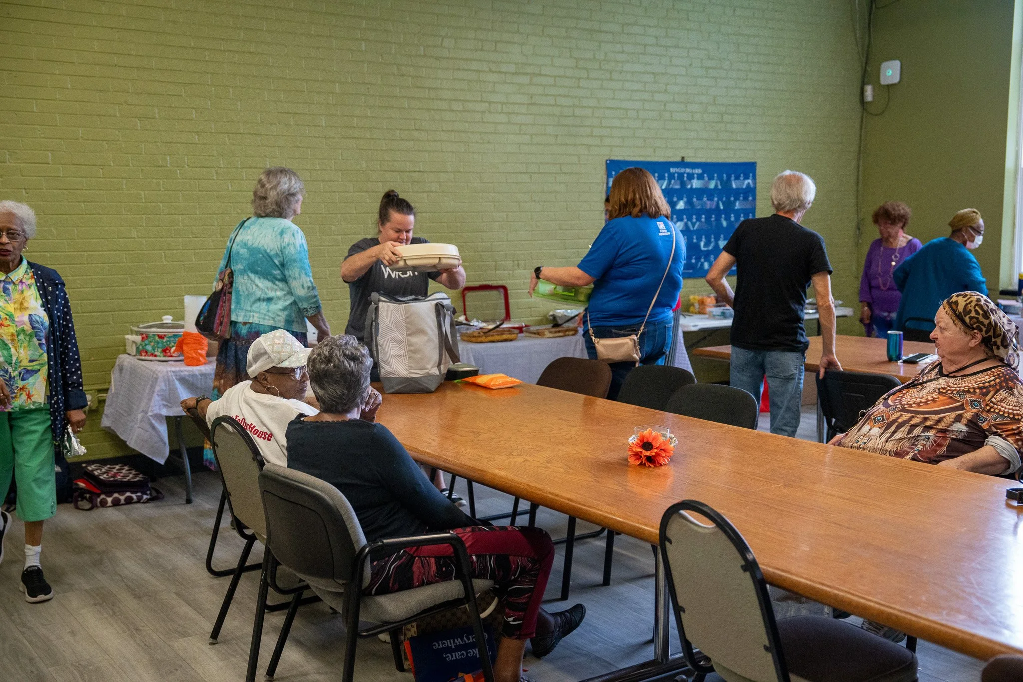 People in a community room with food and supplies, some seated at a long table, others standing near a table with food, in a room with a green brick wall.