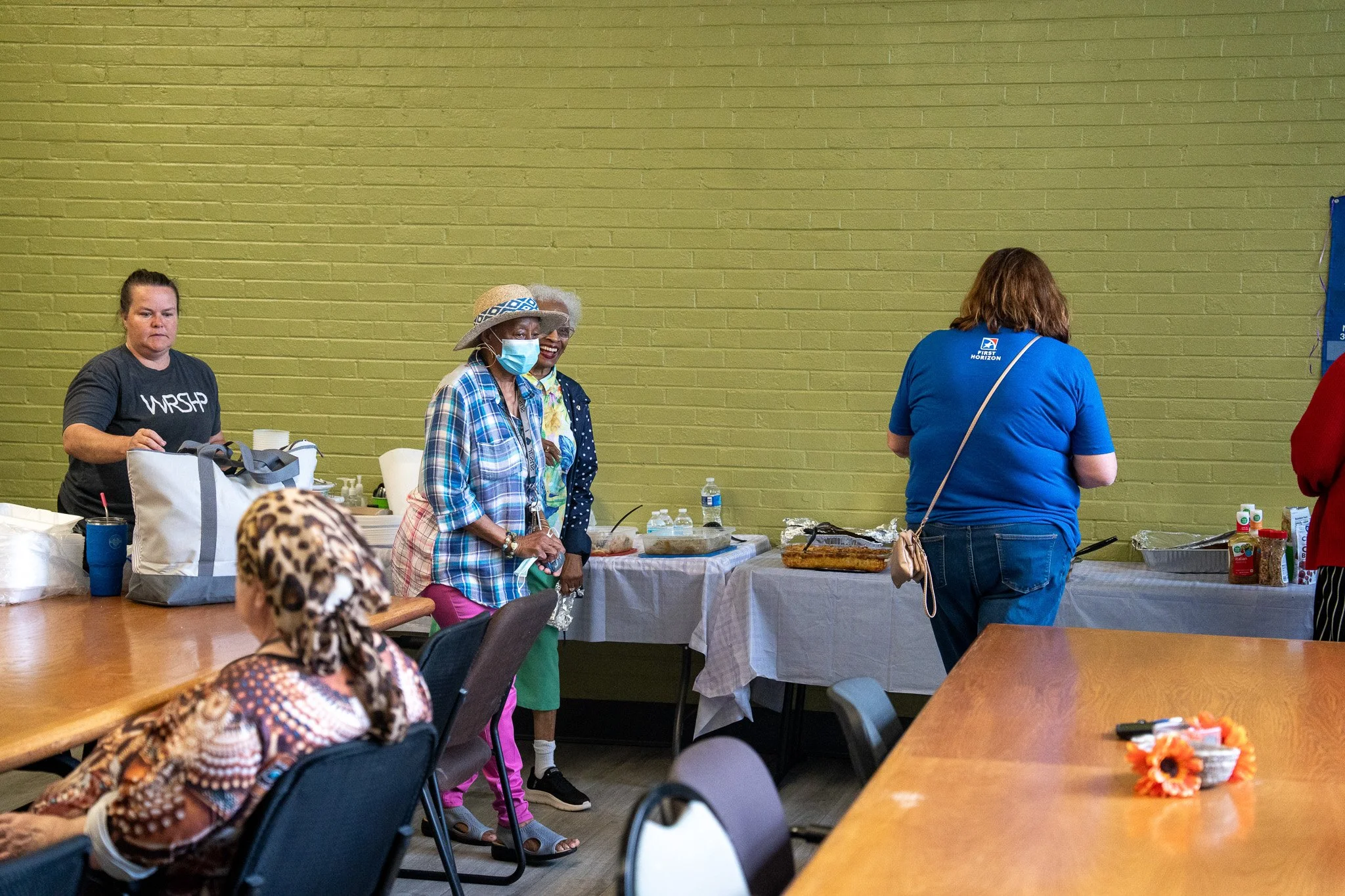 People in a community gathering room with a table of food and a yellow brick wall in the background.