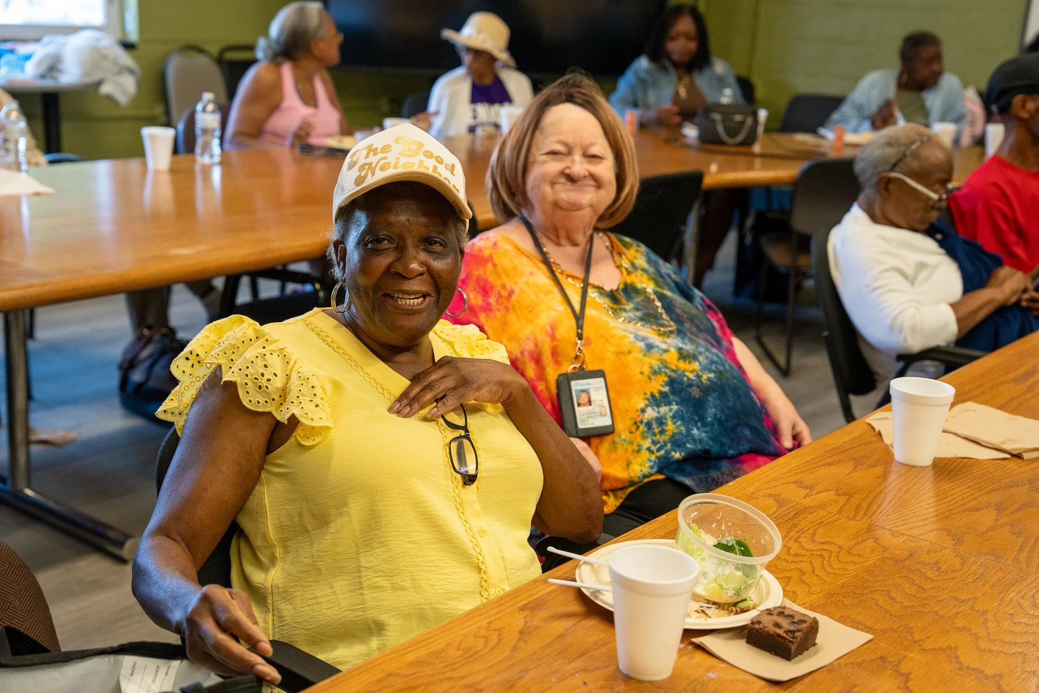 Two smiling women sitting at a table during a gathering or meal, with several other women in the background; one woman is wearing a yellow blouse and a cap, the other is wearing a colorful tie-dye shirt.