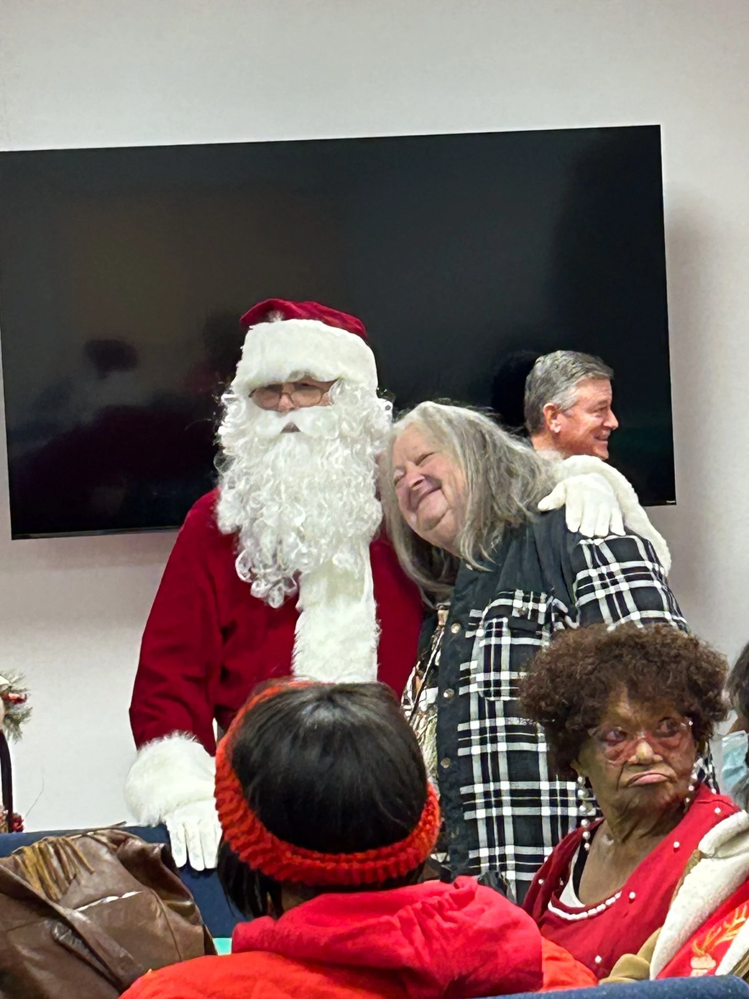 Santa Claus hugging an elderly woman with long gray hair, both smiling warmly. Several other people are visible in the foreground and background.