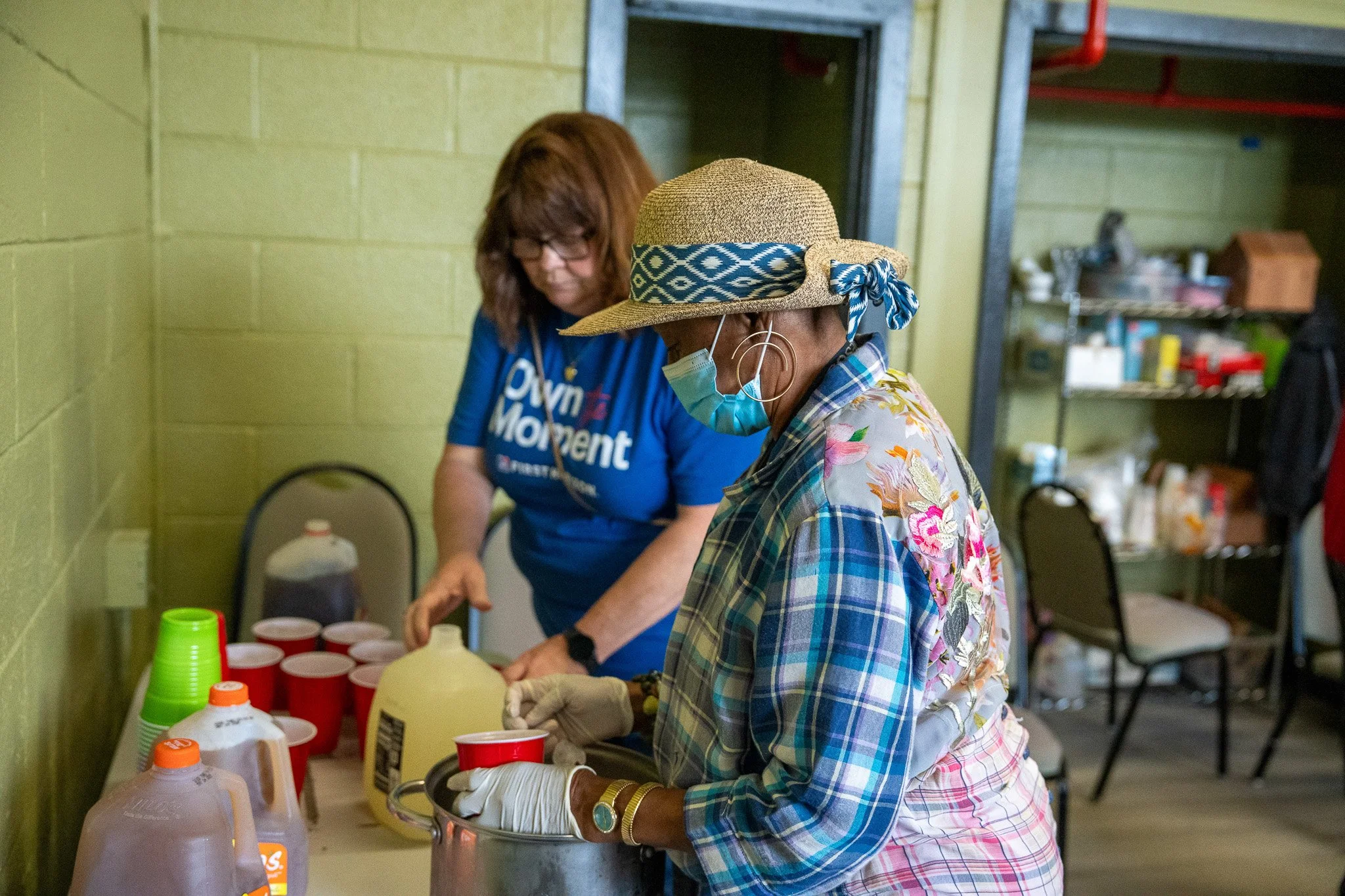 Two women preparing drinks at a table, one wearing a floral shirt, straw hat with a patterned ribbon, and a face mask, the other in a blue campaign shirt, with various drink containers on the table.