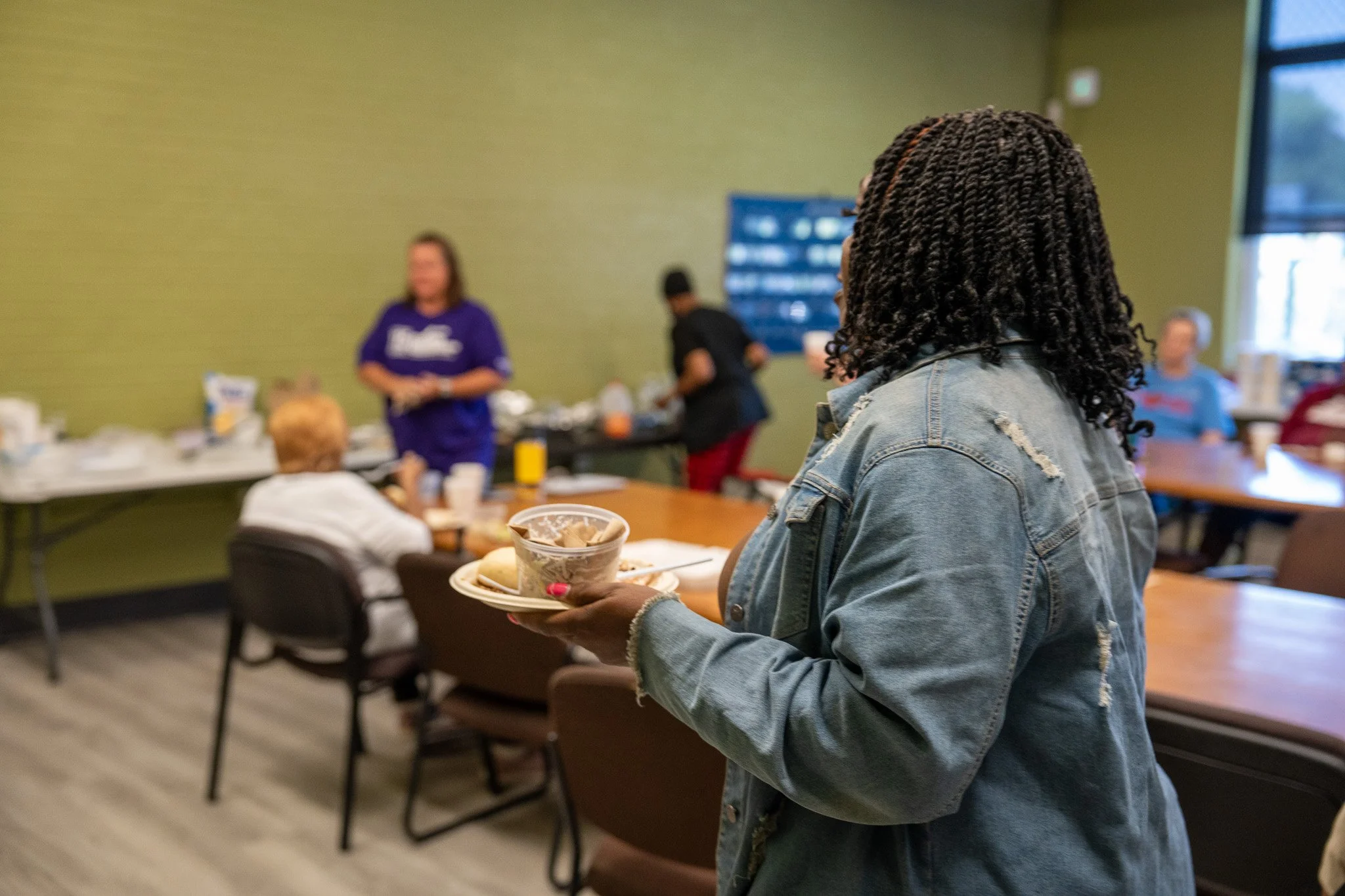 A woman with curly hair wearing a denim jacket holding a plate of food in a community room with other people in the background.