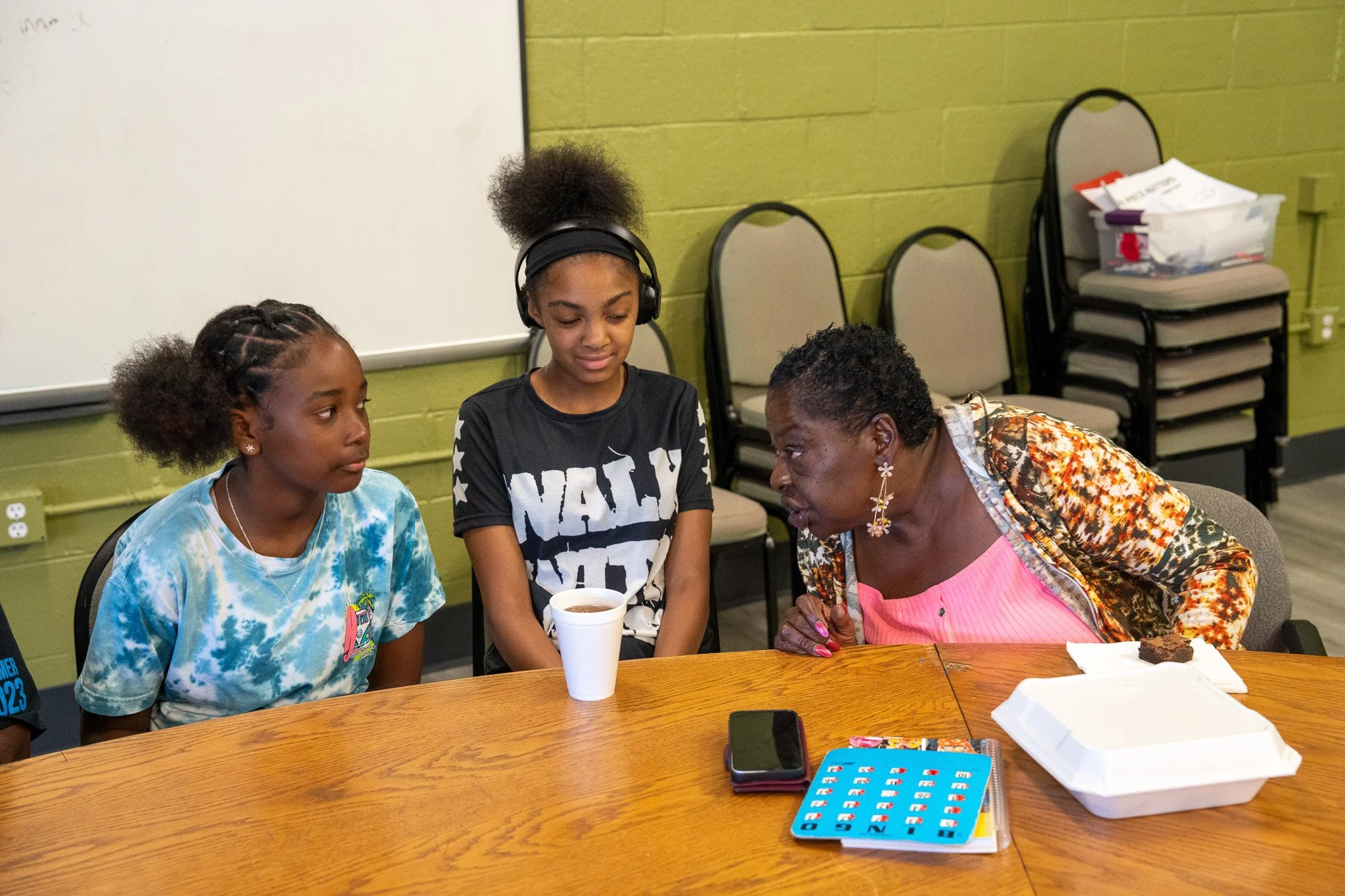 Three women sitting around a table in conversation, one of whom appears to be teaching or explaining something, with a whiteboard and chairs in the background.