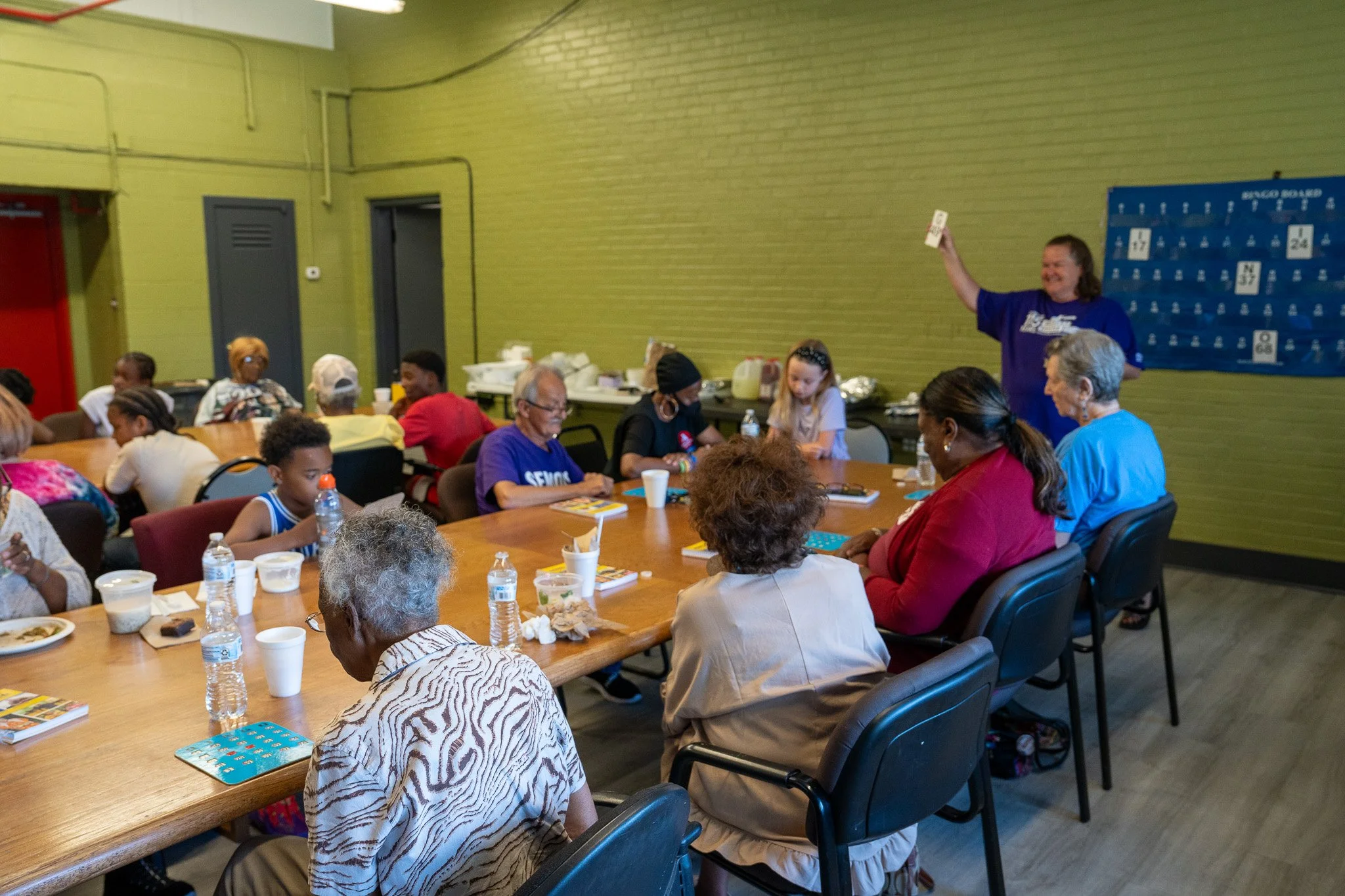 A diverse group of people, including children and elderly, gathered around a large table in a community room. An instructor stands at the front, holding up a card and smiling, as attendees look on attentively. The room has green walls, and there are 