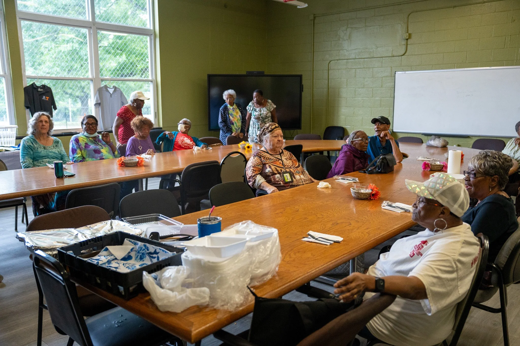 A group of elderly people sitting around a large U-shaped table in a community room, some talking and others listening. The room has large windows with green trees outside, and there is a large TV and a whiteboard on the walls.