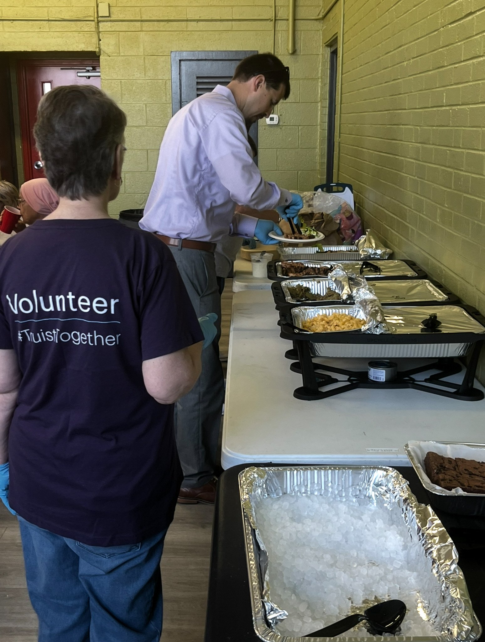 A volunteer in a navy T-shirt with the words '#Twistogether' on the back stands observing a man serving food from a buffet line with trays of different dishes, on a table against a yellow brick wall.