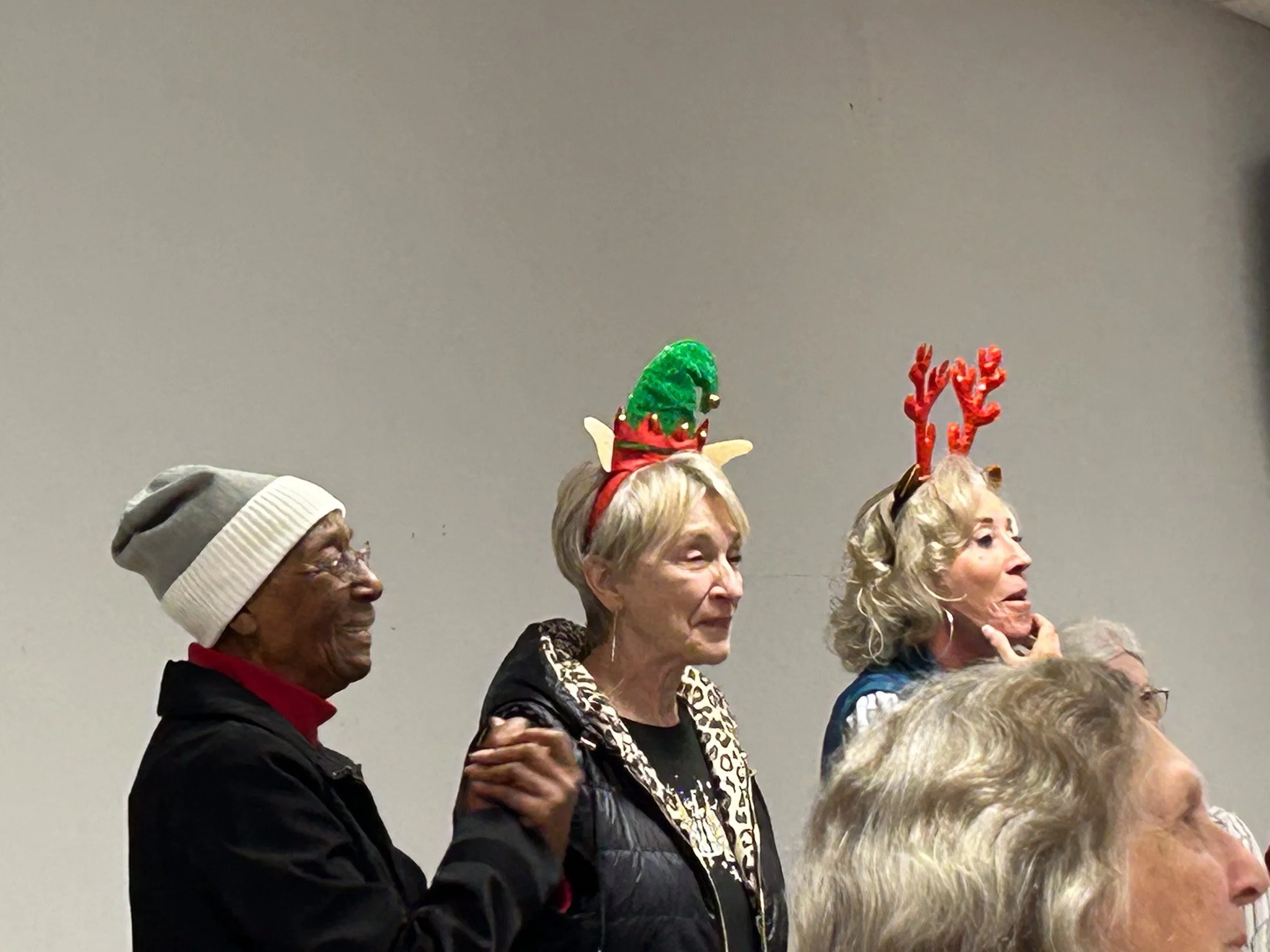 Three women wearing Christmas headbands, standing close together in a room with a plain wall background.