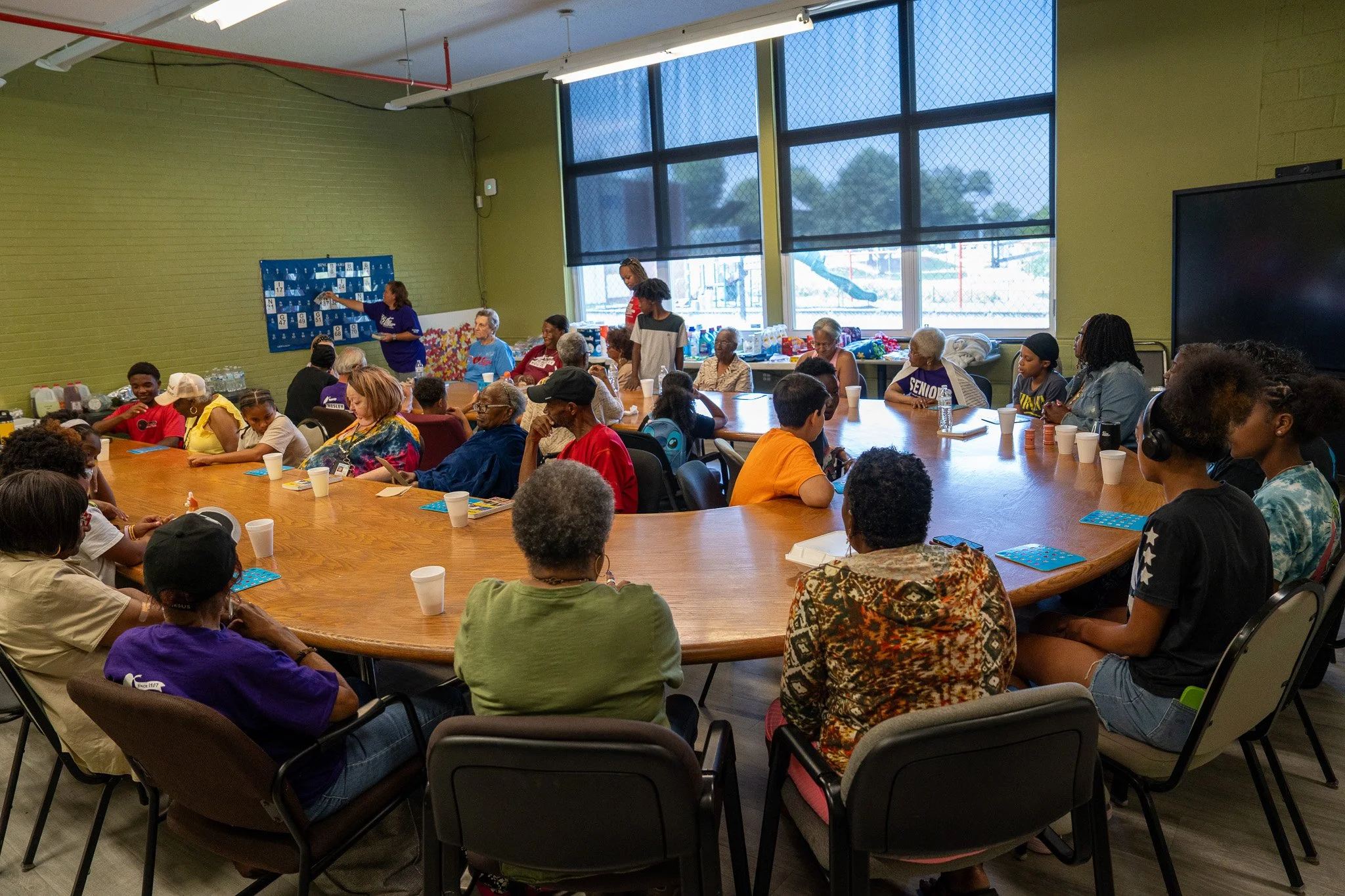 A diverse group of people gathered around a large, oval conference table in a room with green walls and large windows, participating in a meeting or workshop. A woman in a purple shirt is presenting at the front, with various refreshments visible on 