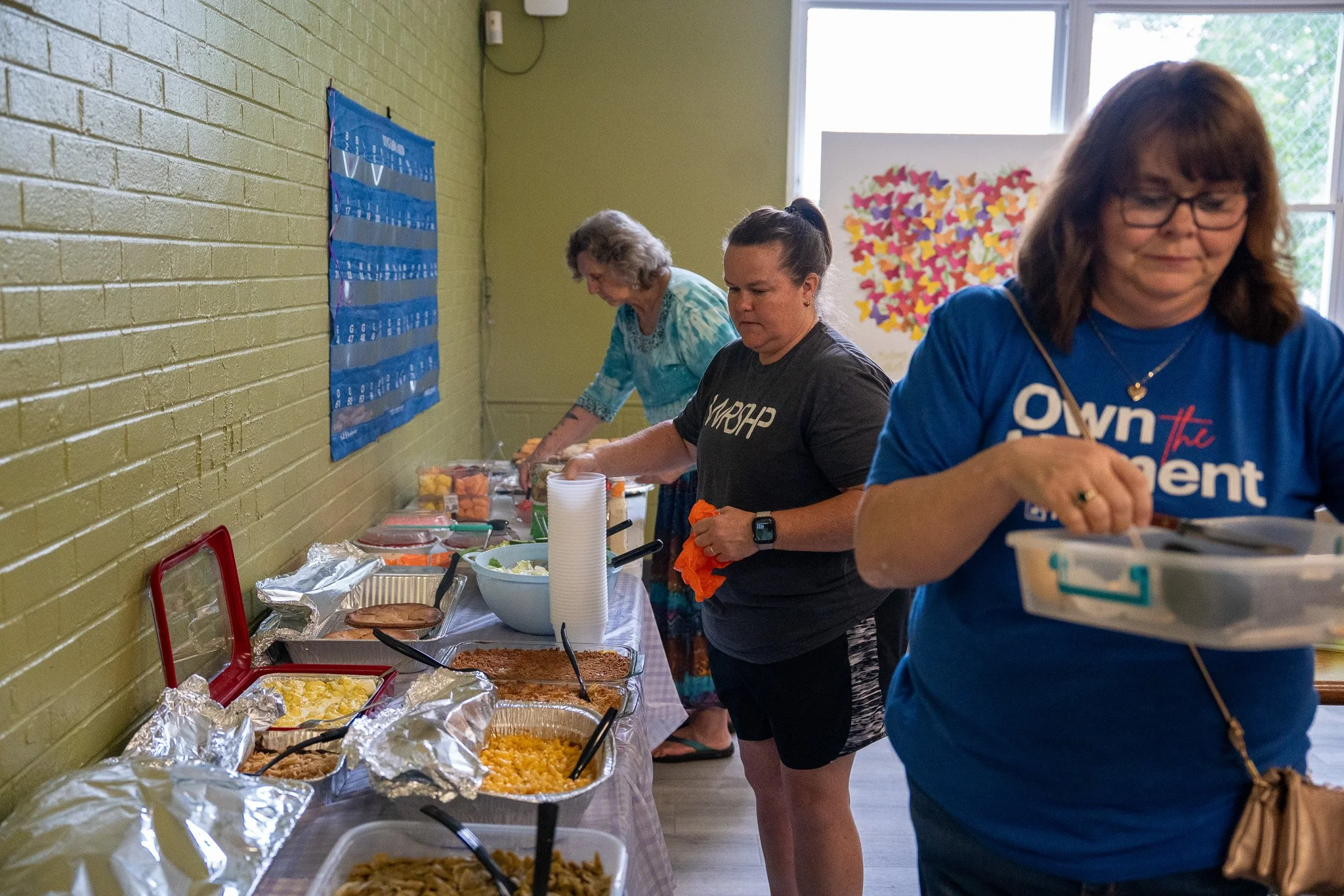 People serving food at a buffet table in a room with yellow brick walls and a window, with a colorful butterfly artwork in the background.