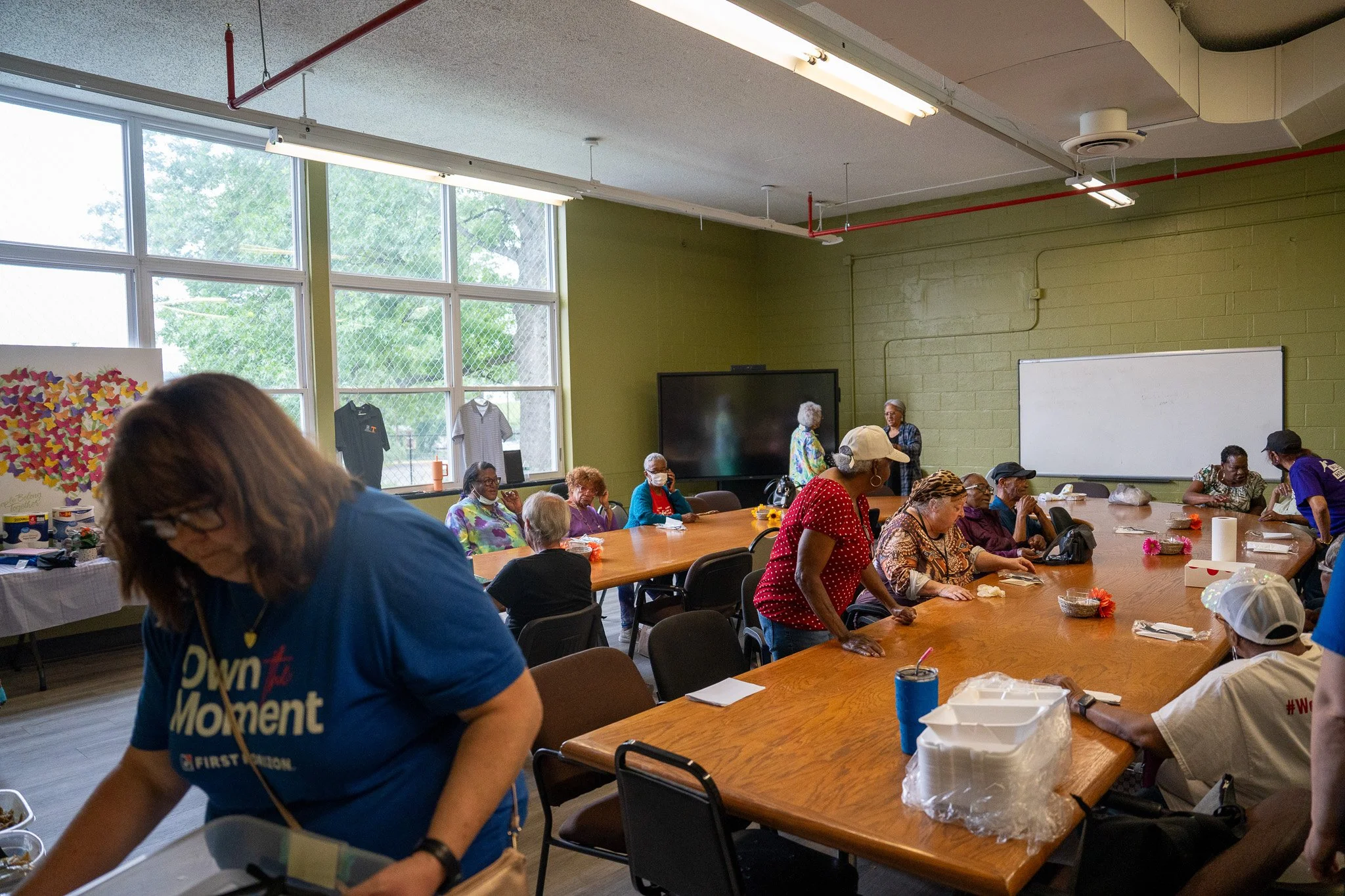 People gathered in a classroom-style room with large windows, some sitting at a long table, others standing and talking, with whiteboard and large screen at the front.