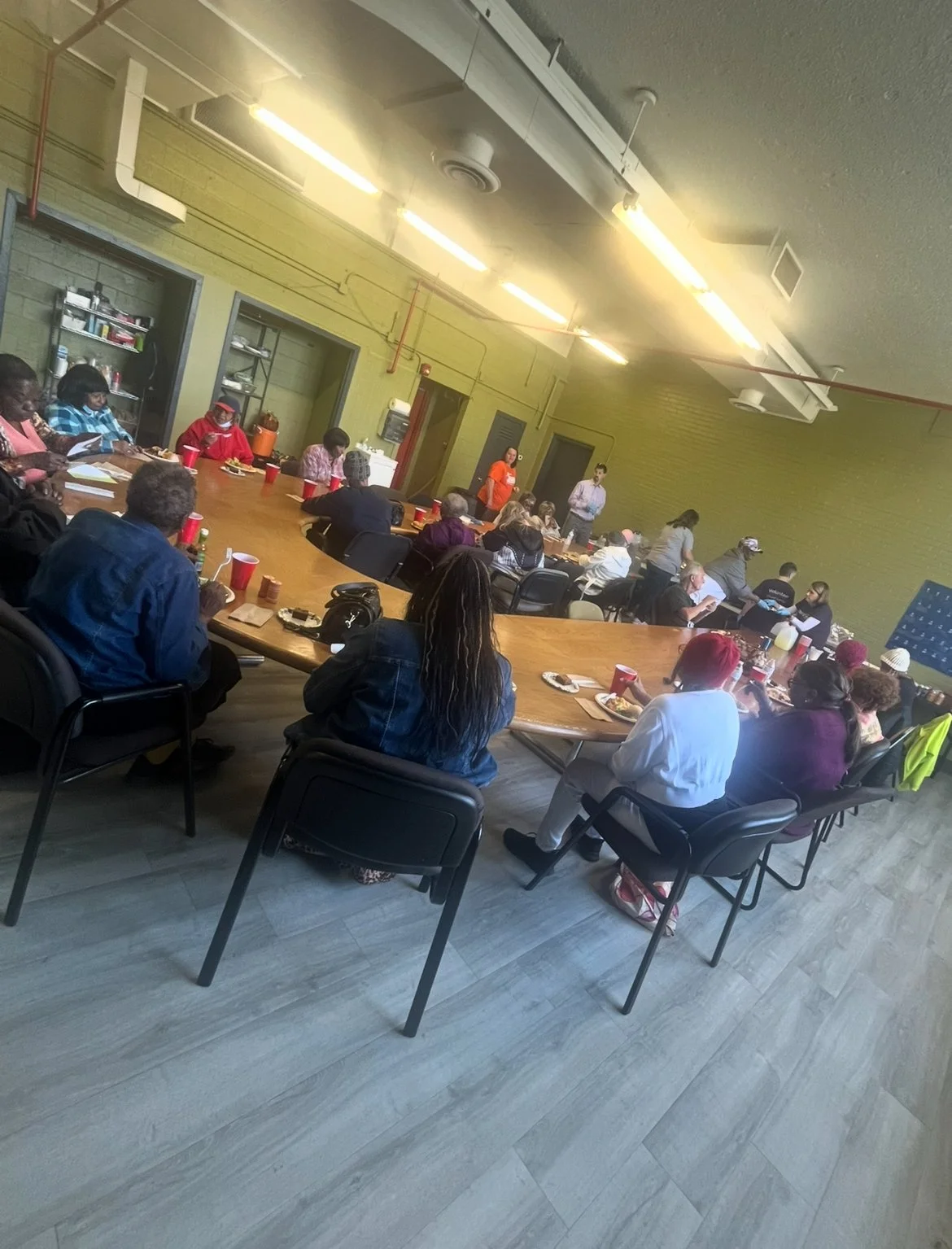A group of people seated around a large conference table in a meeting room, with some individuals at the front near a whiteboard and a person speaking.