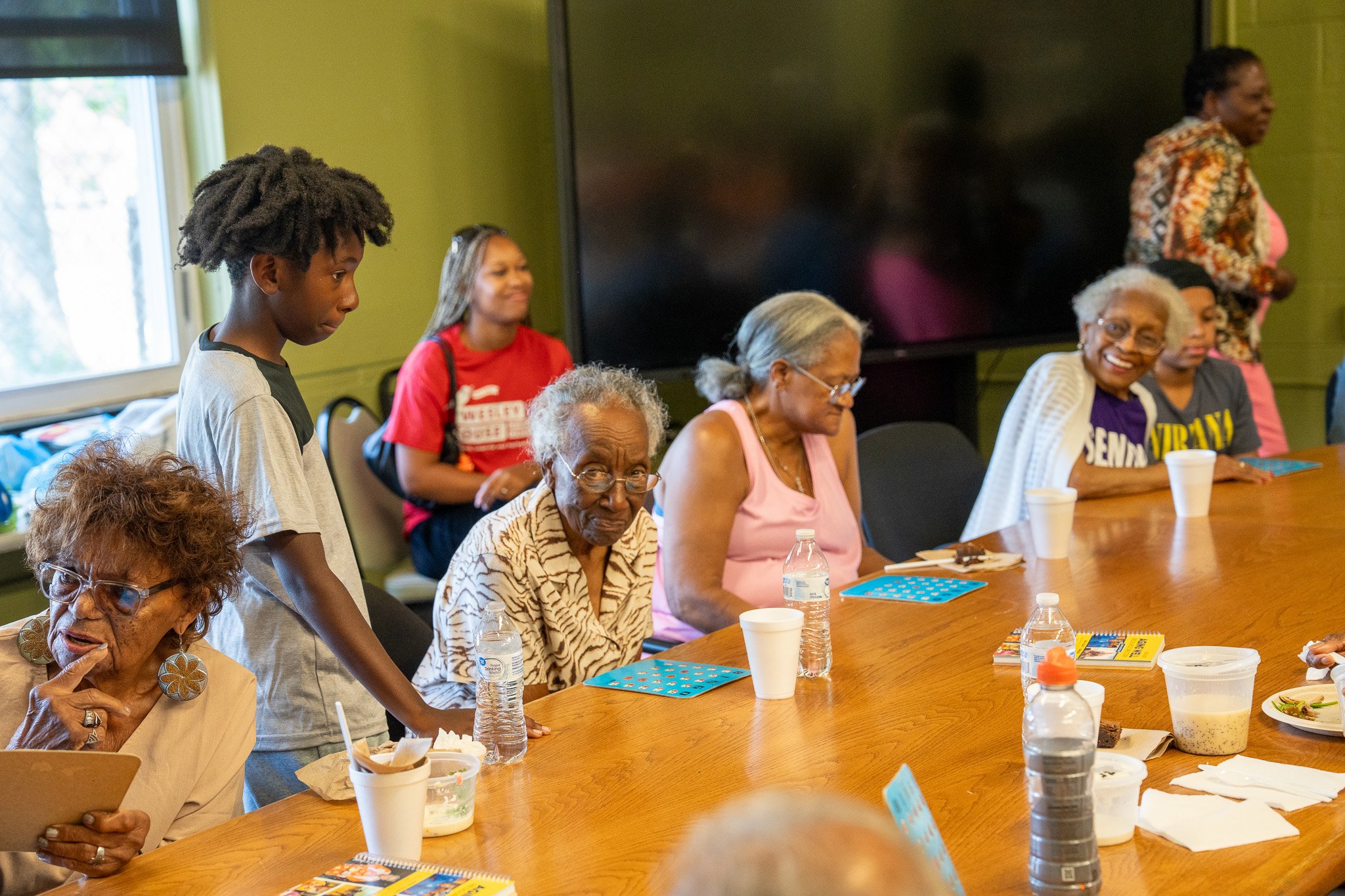 A group of elderly women and a young girl gathered around a conference table, some sitting and some standing, with snacks and drinks on the table in a room with green walls and a large window.