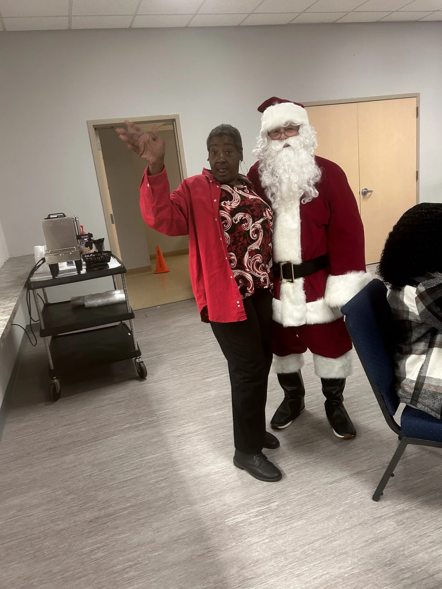 A woman standing next to a person dressed as Santa Claus, who is waving and smiling, inside a room with a beige wall and door.