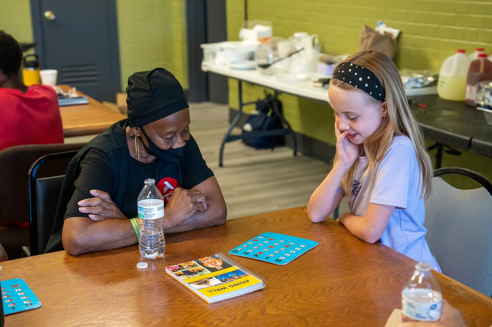A woman and a young girl playing a game at a wooden table in a room with green walls and a table with food and drinks in the background.