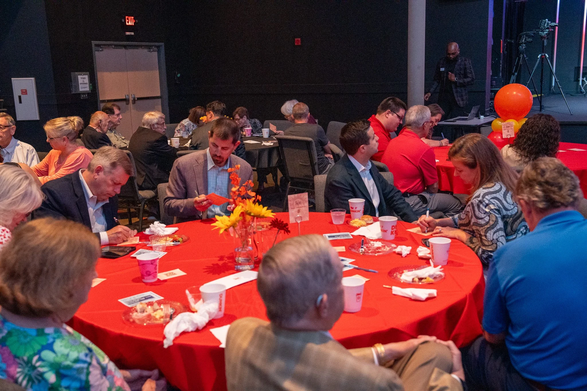 People seated at round tables with red tablecloths, eating and engaging in activities at an indoor event, with a stage and cameraman in the background.