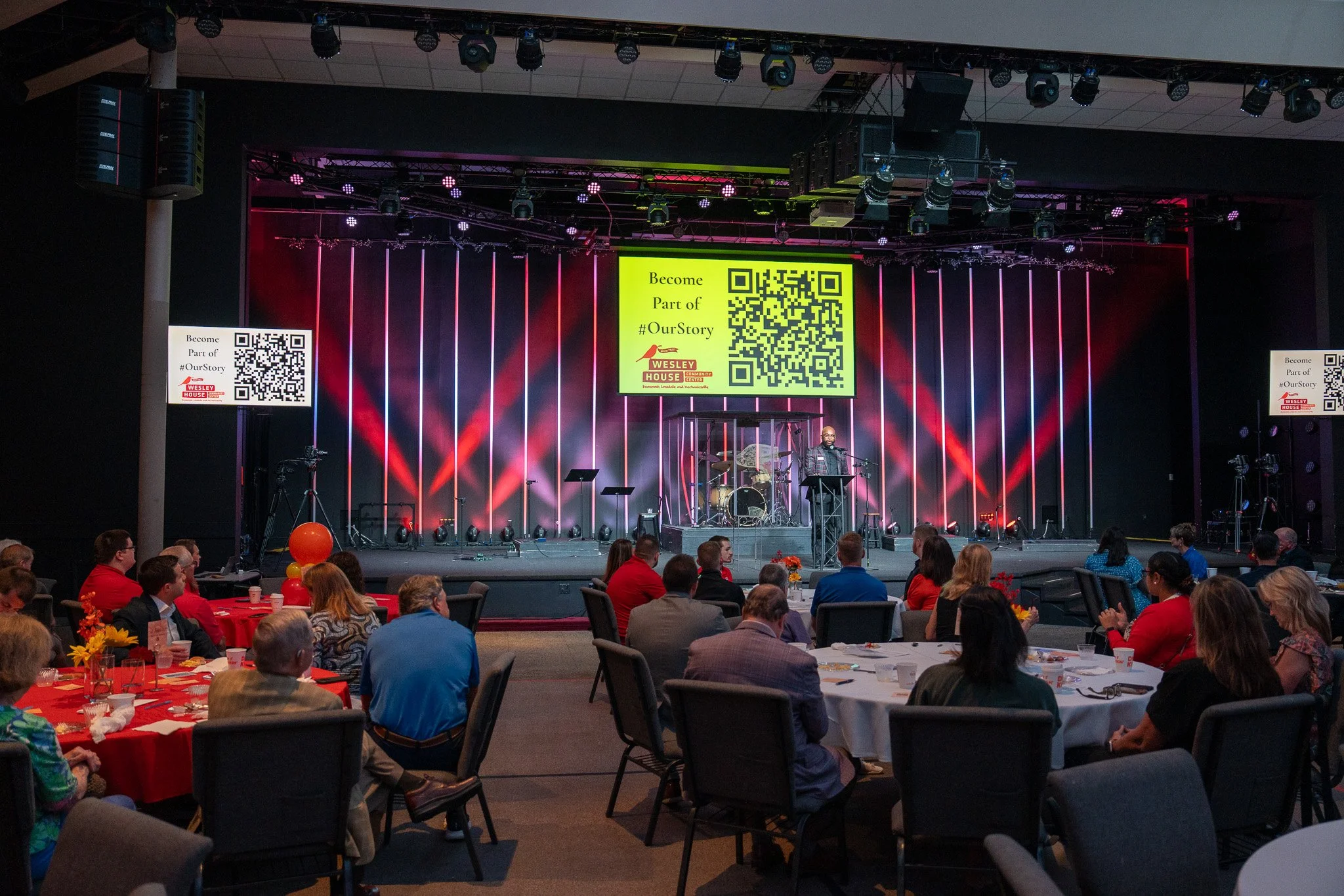 A gathering seated at round tables with red tablecloths in a large hall, facing a stage illuminated with multicolored lights. On the stage, a speaker stands at a podium, with a drum set behind him. Large screens above display a QR code and text encou
