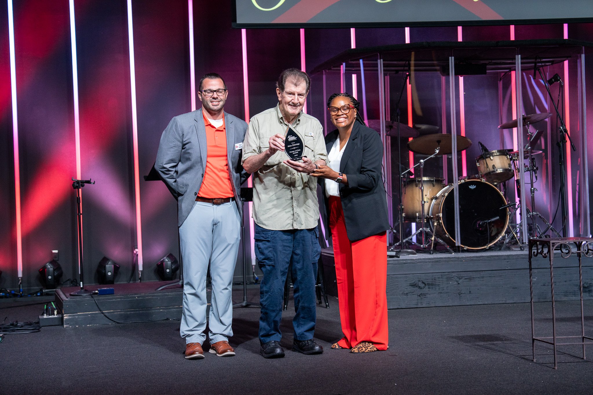 Three people on a stage holding an award plaque. The background shows a drum set and stage lights.