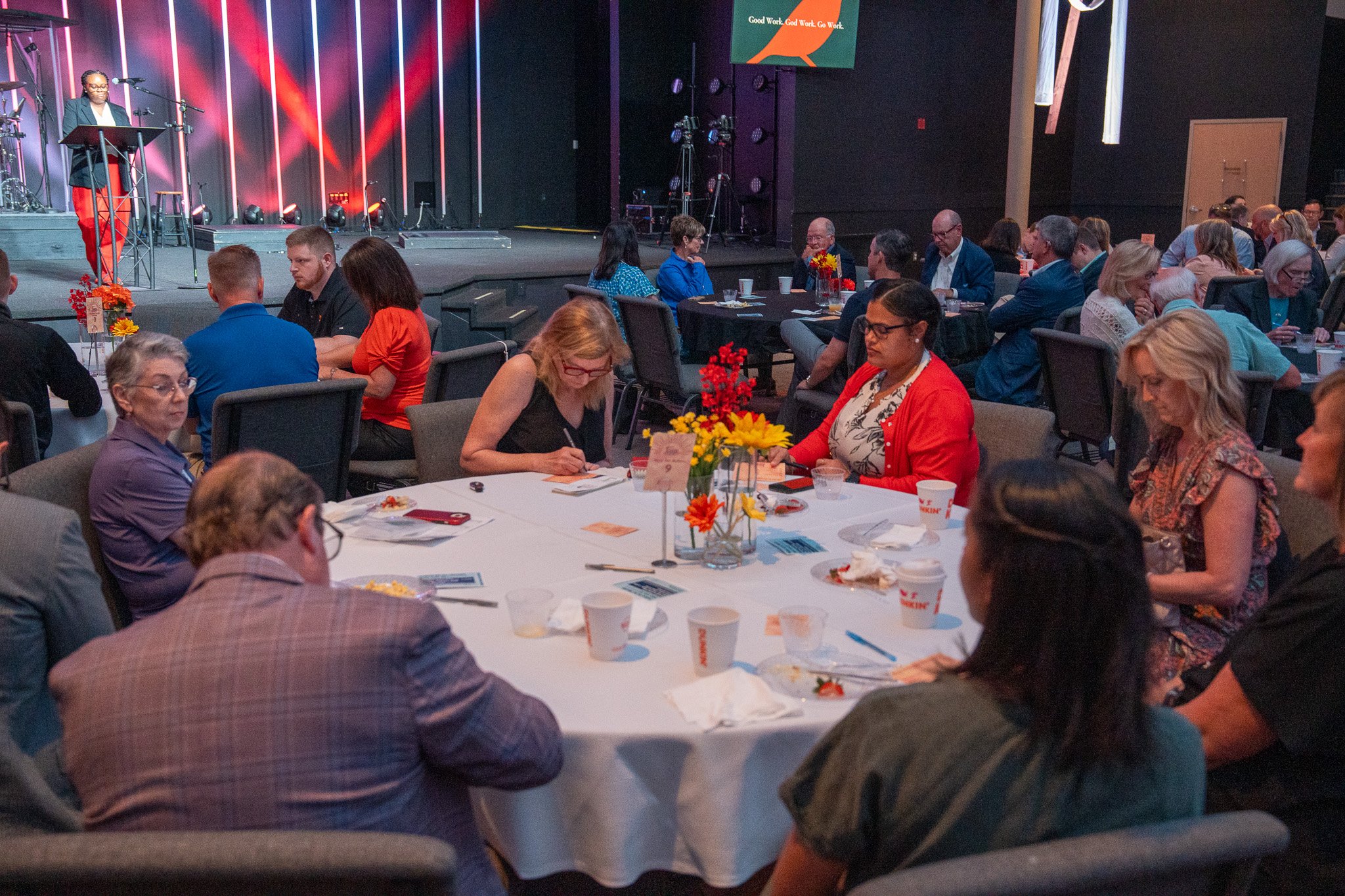 People sitting around a round banquet table at an indoor event, some taking notes, with a stage and speaker in the background.