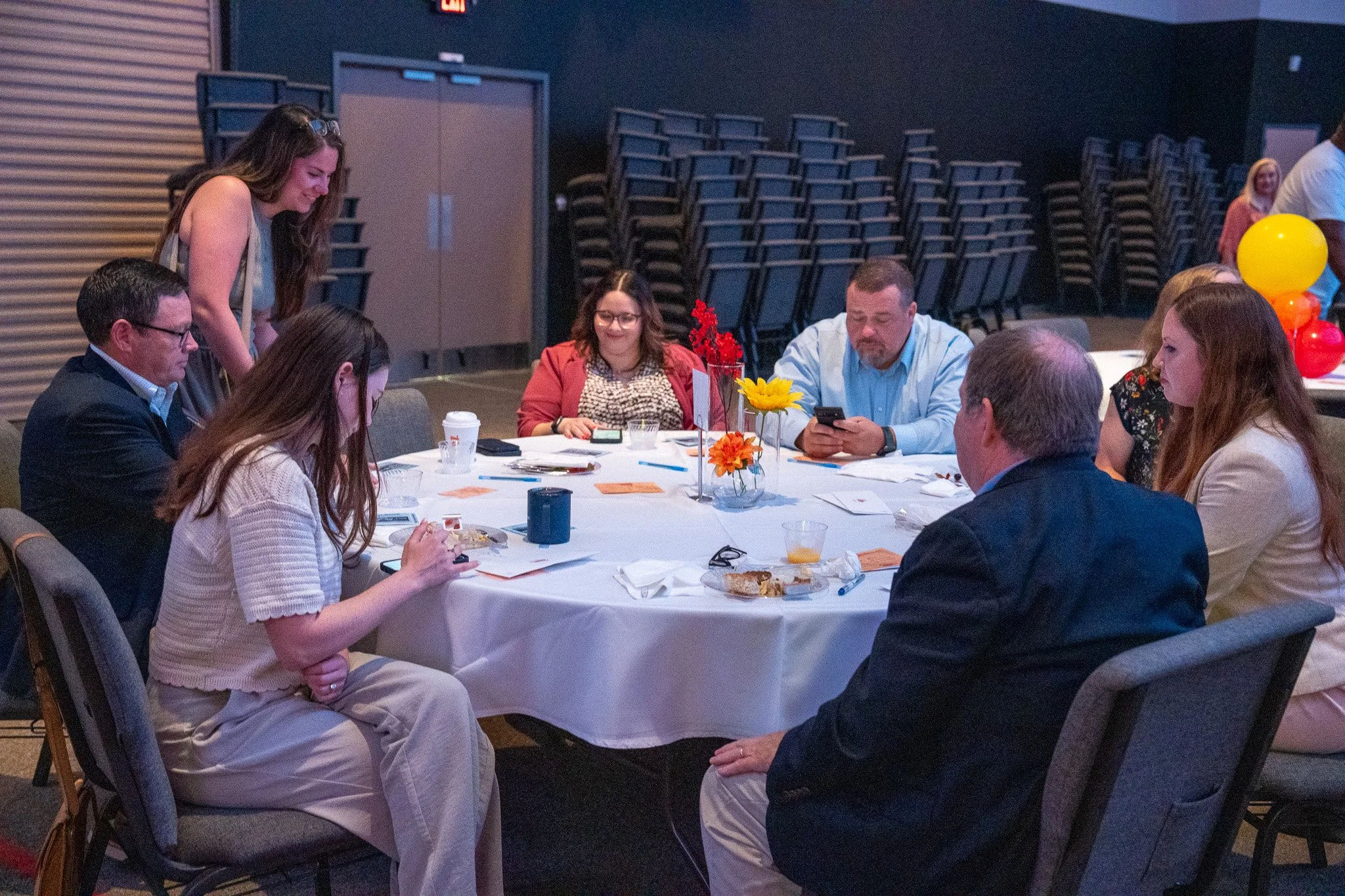 Group of people sitting around a banquet table at an indoor event, some looking at their phones, with balloons and flowers in the background.