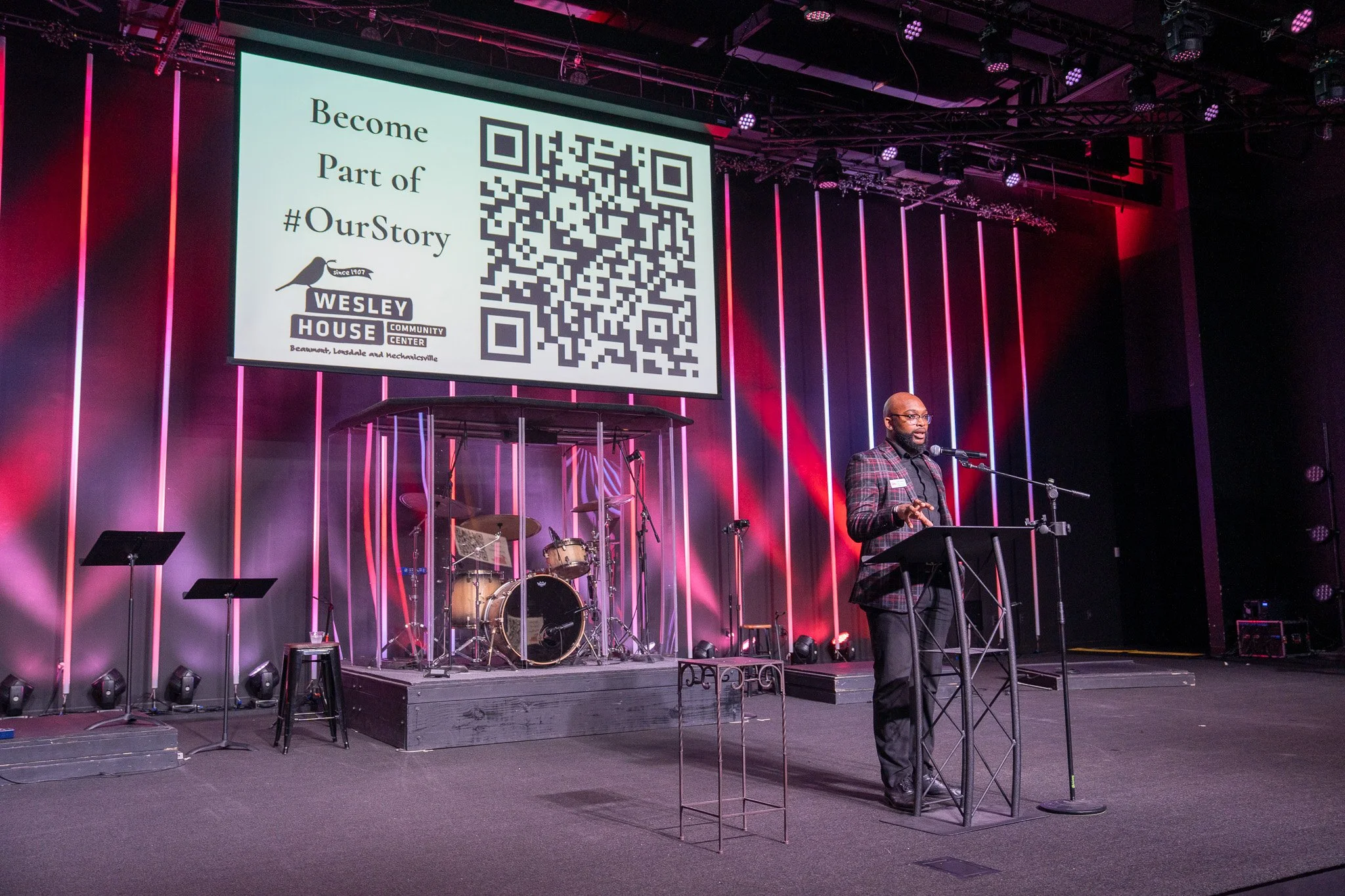 A man in a plaid blazer stands at a podium with a microphone, speaking at an event with a large screen behind him. The screen displays a QR code, the Wesley House Community Center logo, and the message 'Become Part of #OurStory.' The stage has musica