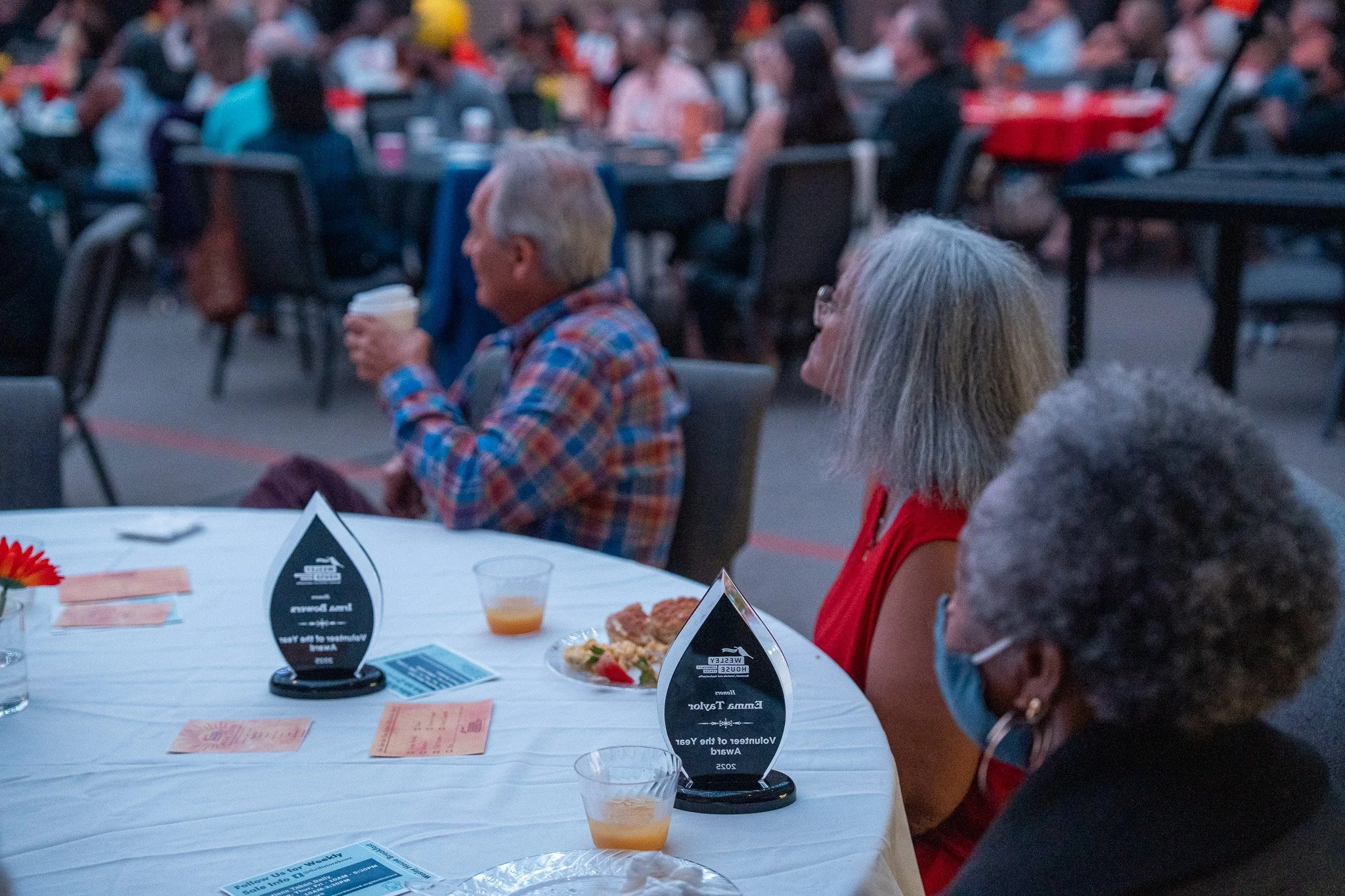 People sitting at a round table with awards, food, and drinks, at an outdoor event during evening or dusk. Some individuals are wearing masks.