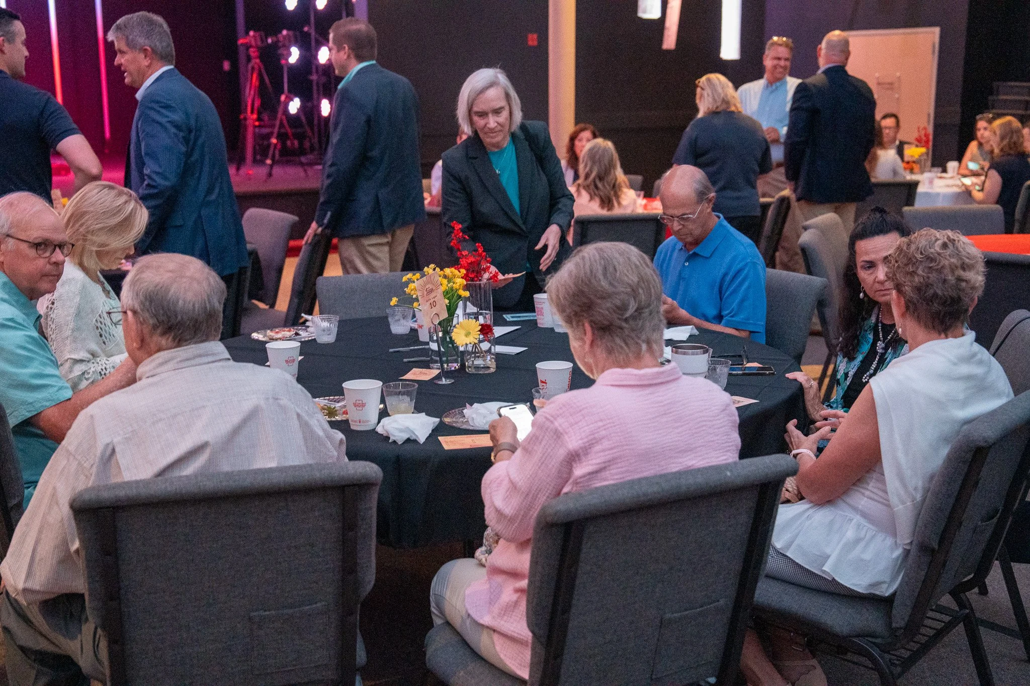 People sitting around a table at a social event, with some standing and chatting in the background. The table has floral centerpieces and food, with a stage and lighting in the background.