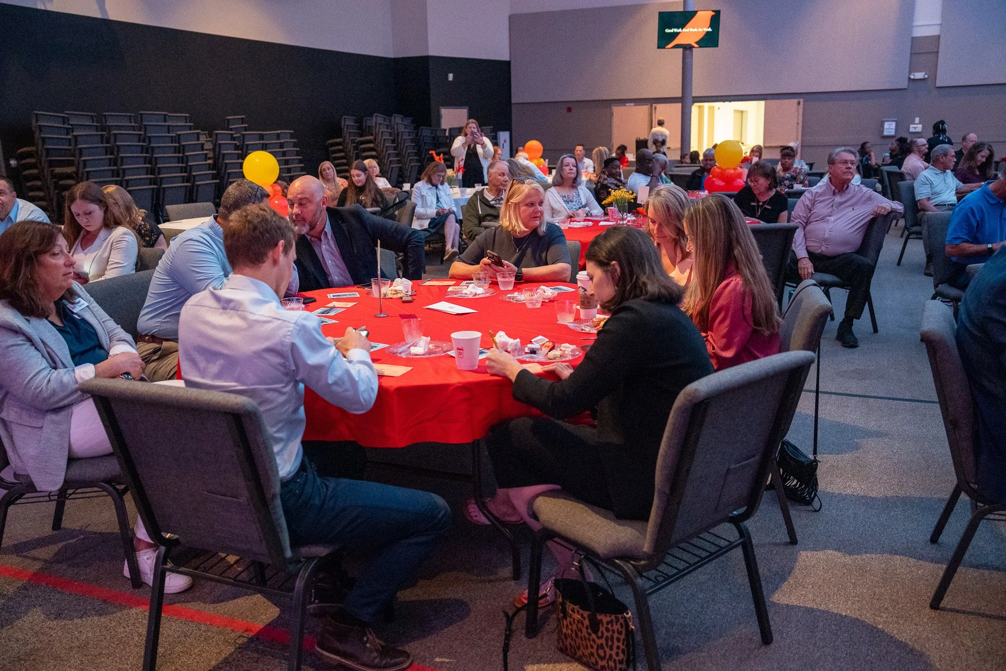 People gathered around tables with red tablecloths at an indoor event or conference, some are eating, talking, or using their phones. Balloons and flowers decorate the tables, and a large screen is visible in the background.