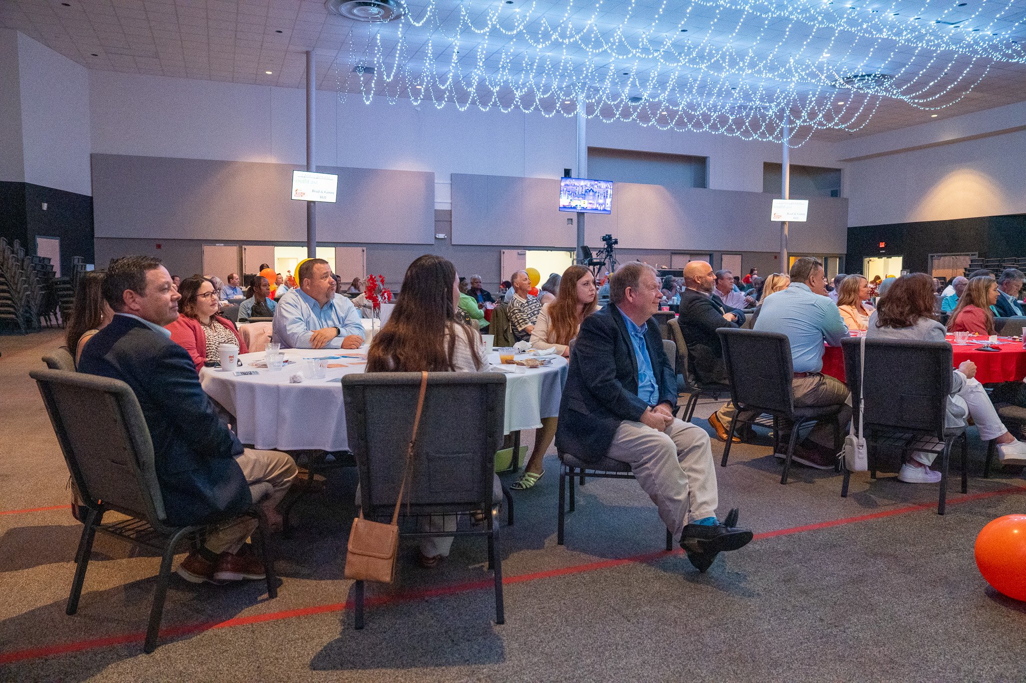 Audience attending a conference or event in a large indoor hall, seated at round tables covered with white tablecloths, with some tables having red coverings. The hall has a high ceiling decorated with string lights. Multiple television screens are m