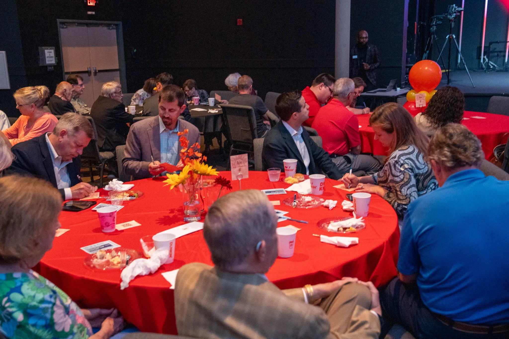 People sitting at decorated round tables with red tablecloths, eating and writing notes, at an indoor event or celebration.