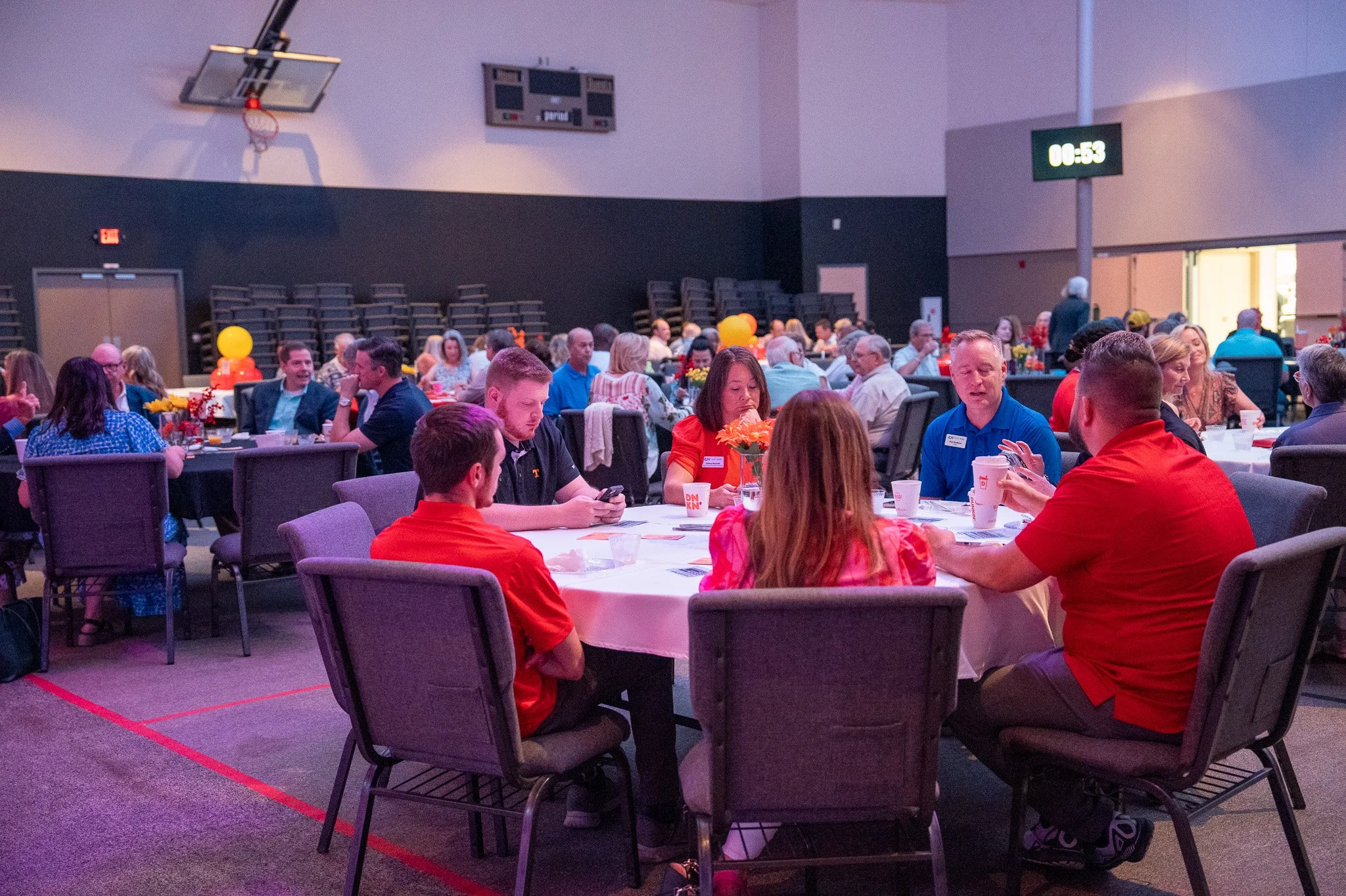 People sitting at round tables in a large event hall with decorated tables, balloons, and a digital clock on the wall, during a social or community event.