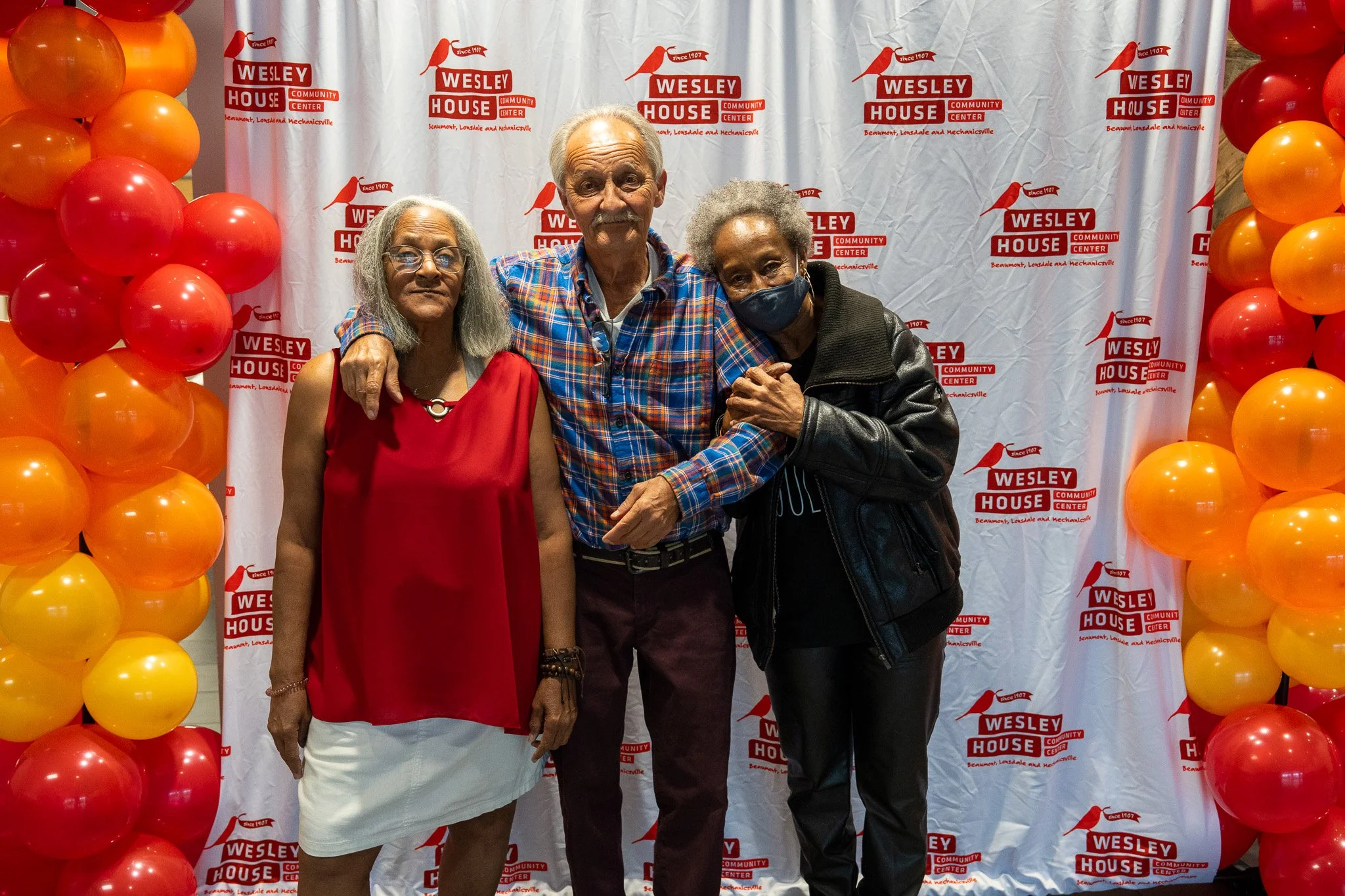 Three people posing together in front of a Wesley House Community Center backdrop with red and orange balloons on either side.