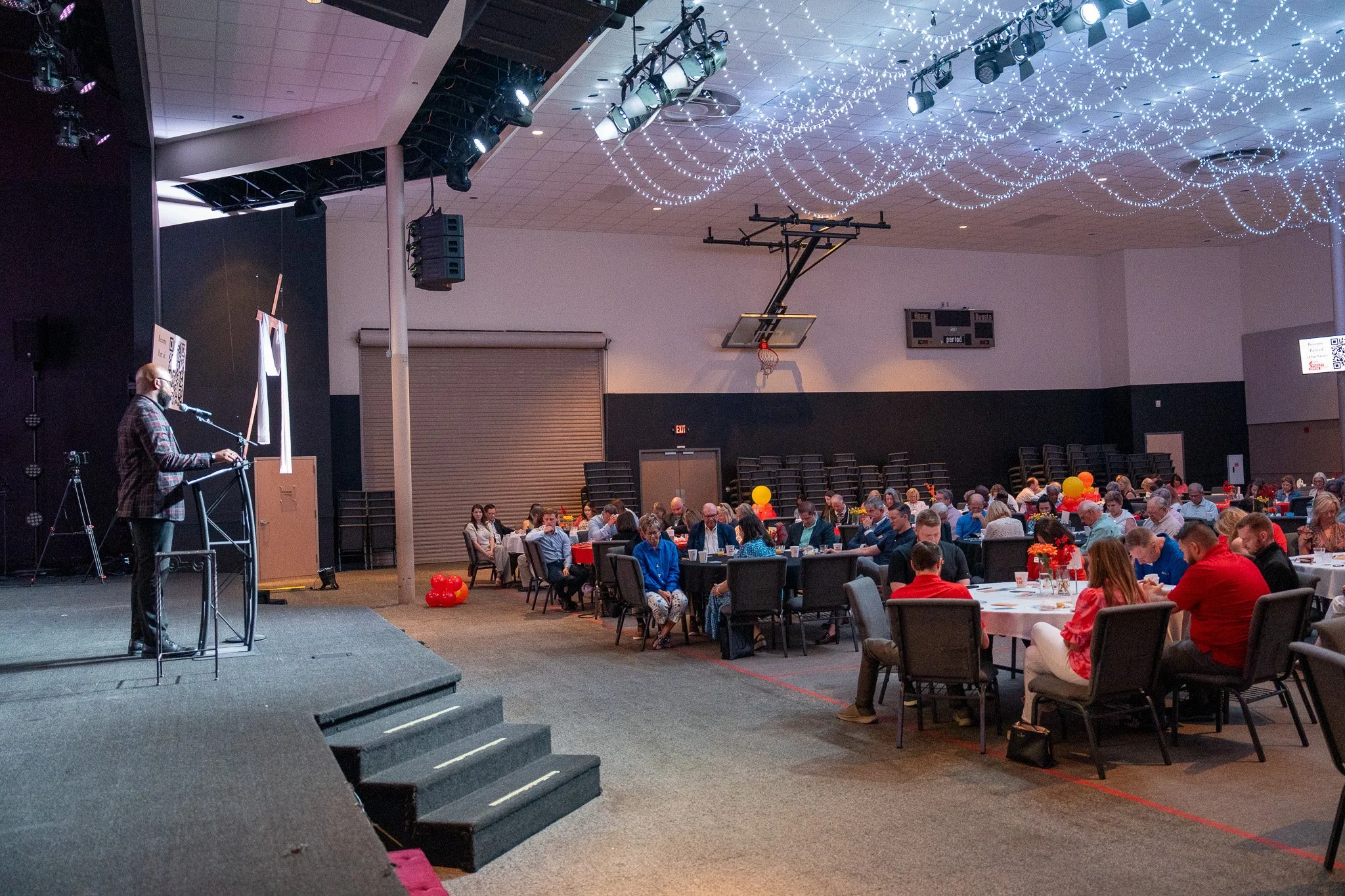 An indoor event with a stage on the left side and numerous round tables filled with people. The stage has a person speaking into a microphone, with a camera on a tripod nearby. The ceiling is decorated with string lights, and there are balloons and f