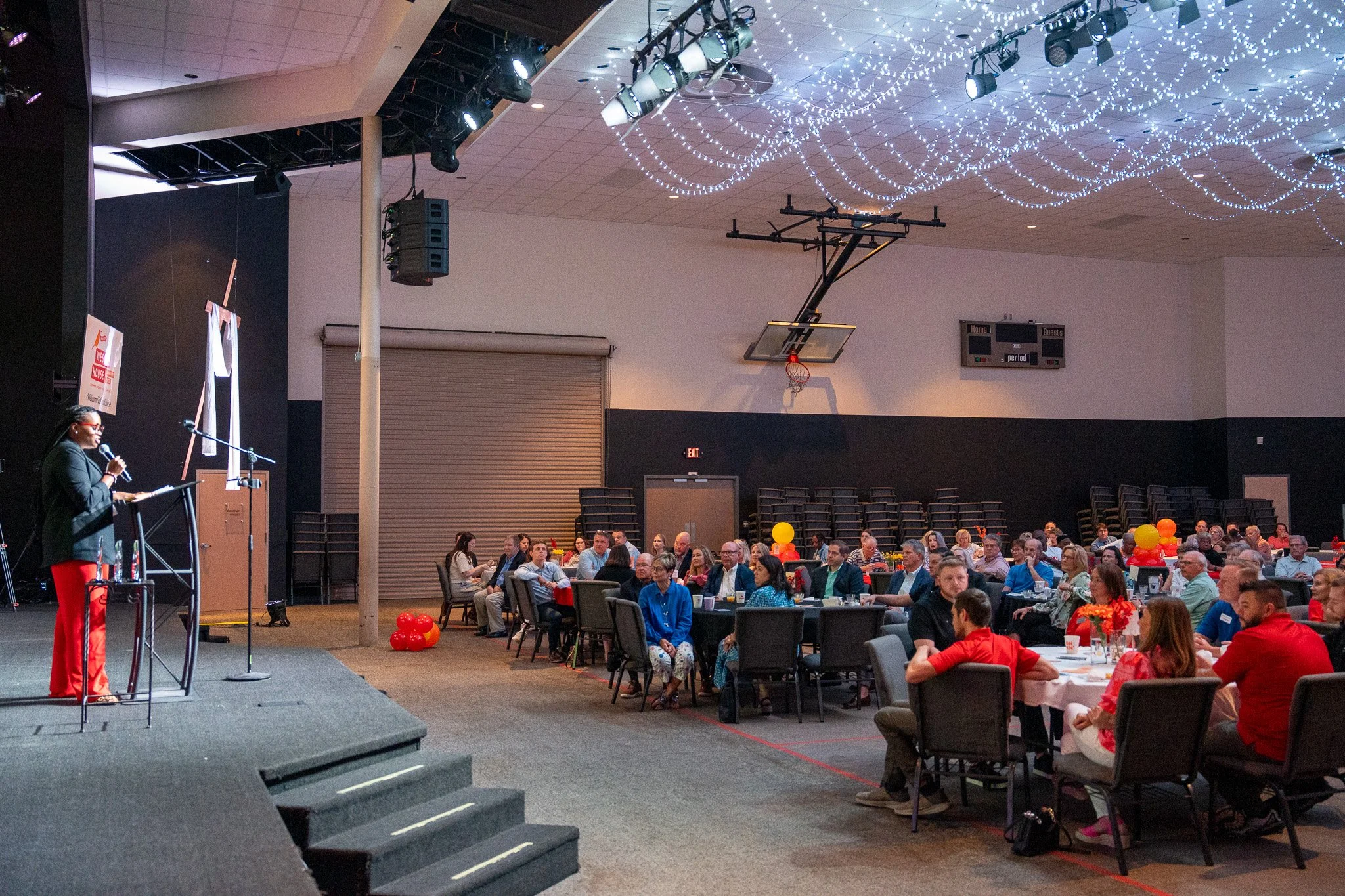 A woman speaking at a podium in front of an audience seated at round tables in a large decorated event hall, with balloons and string lights overhead.