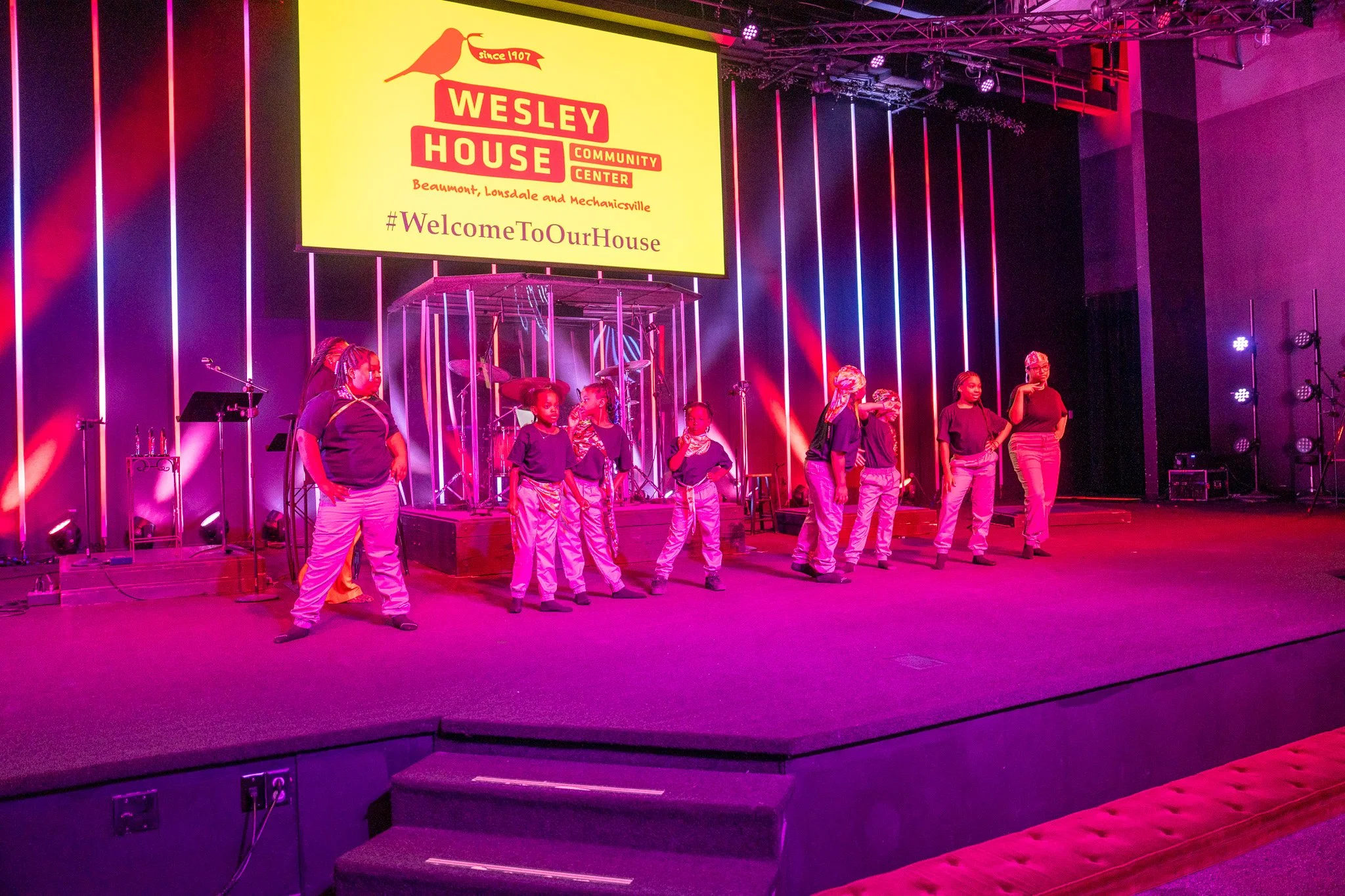 Children performing on a stage with colorful lighting, in front of a large yellow screen displaying the Wesley House Community Center logo and hashtag.