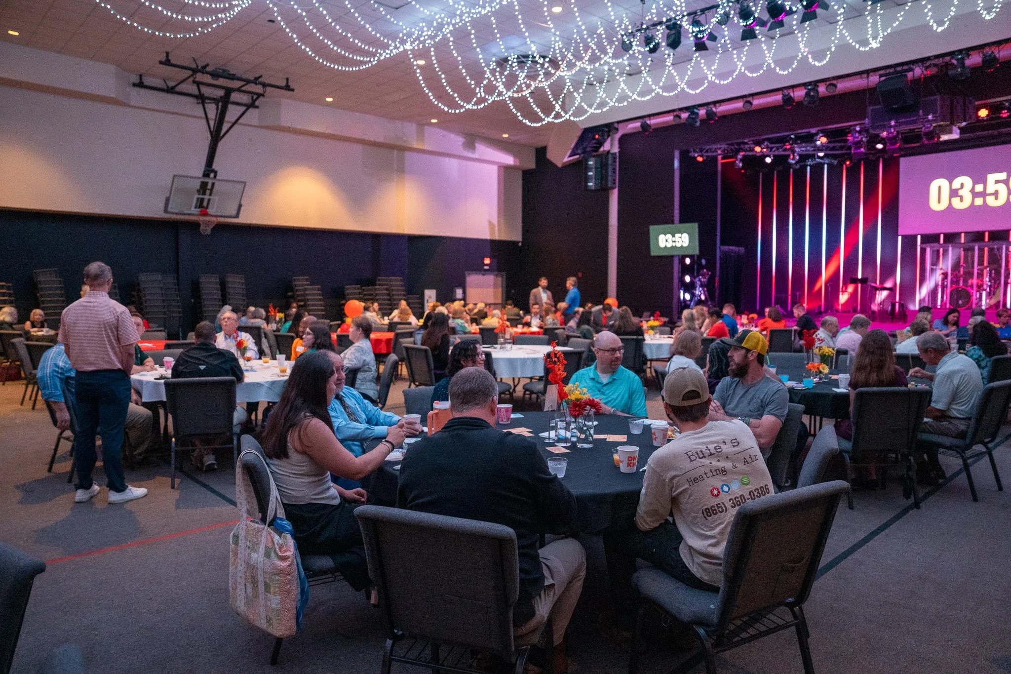 People seated at round tables with orange and yellow flower centerpieces in a large event hall with a stage, decorated with string lights hanging from the ceiling, and a countdown timer on screen.