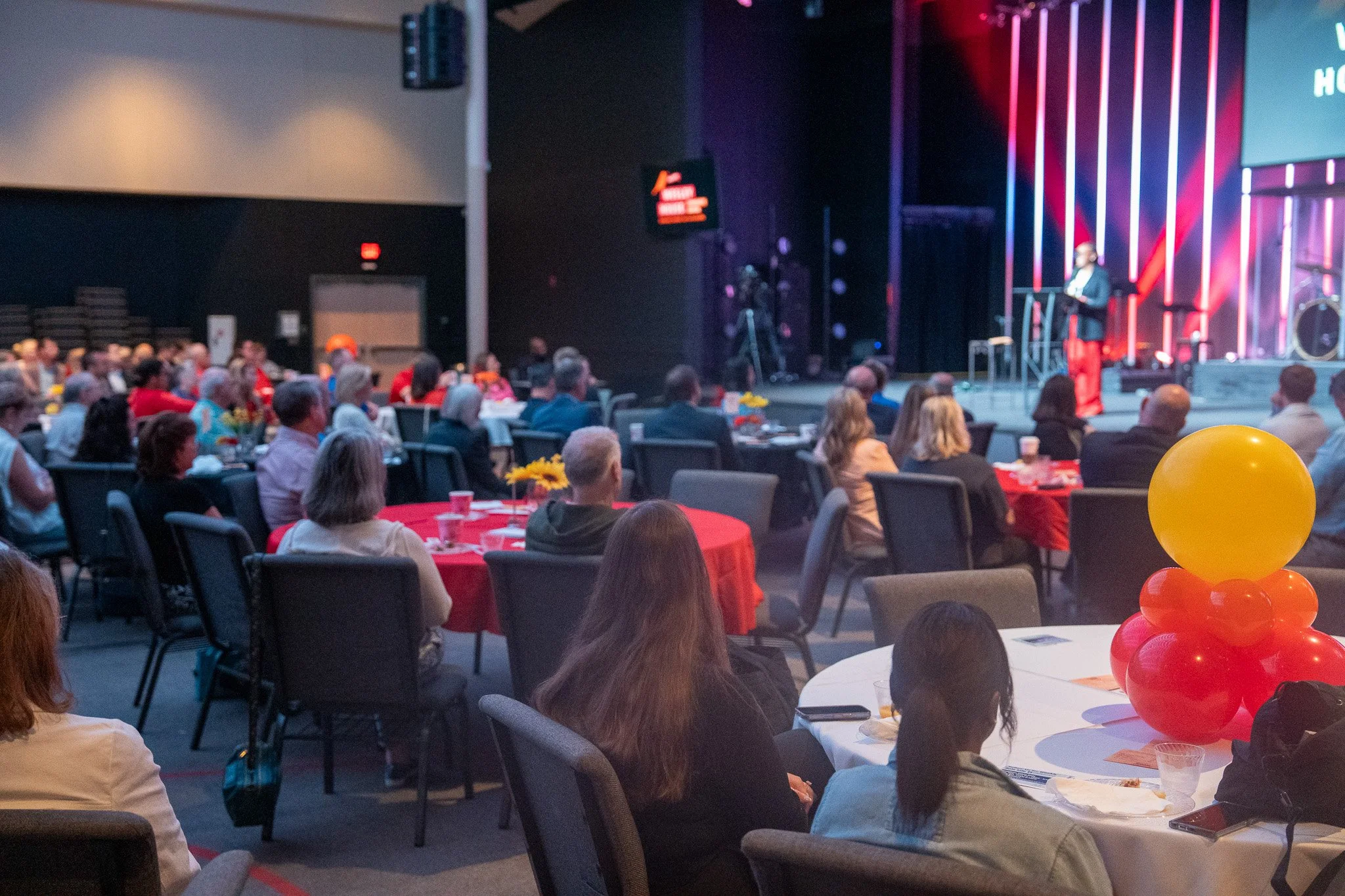 People seated at round tables with red and white tablecloths in a large indoor venue, watching a speaker on stage. The stage has a purple and red lighting backdrop, and balloons are visible on one table.