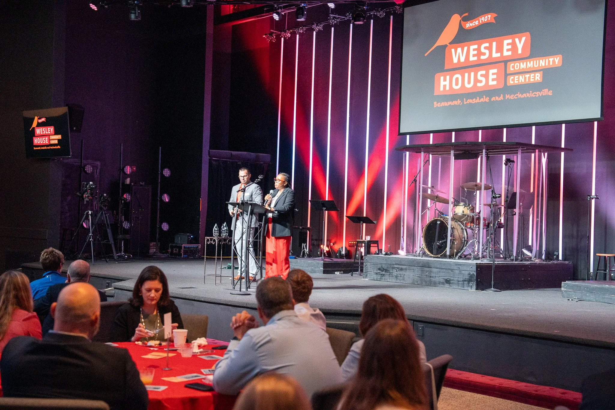 Speakers on stage at Wesley House Community Center event, with a large screen displaying logo and name, audience seated at tables, and stage lighting.