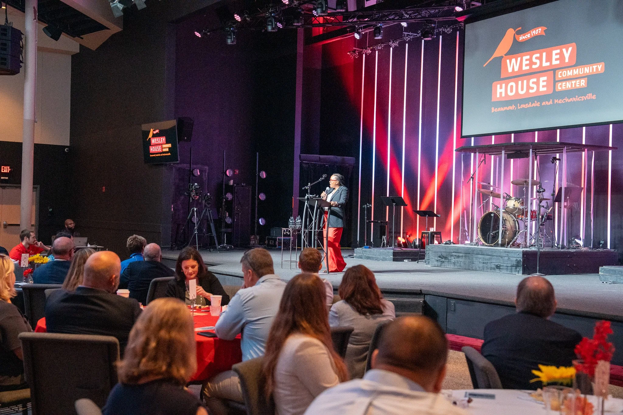 A speaker stands at a podium on stage at the Wesley House Community Center event, with a band setup behind them, in a large auditorium filled with seated attendees listening to the presentation, with screens displaying the Wesley House logo.