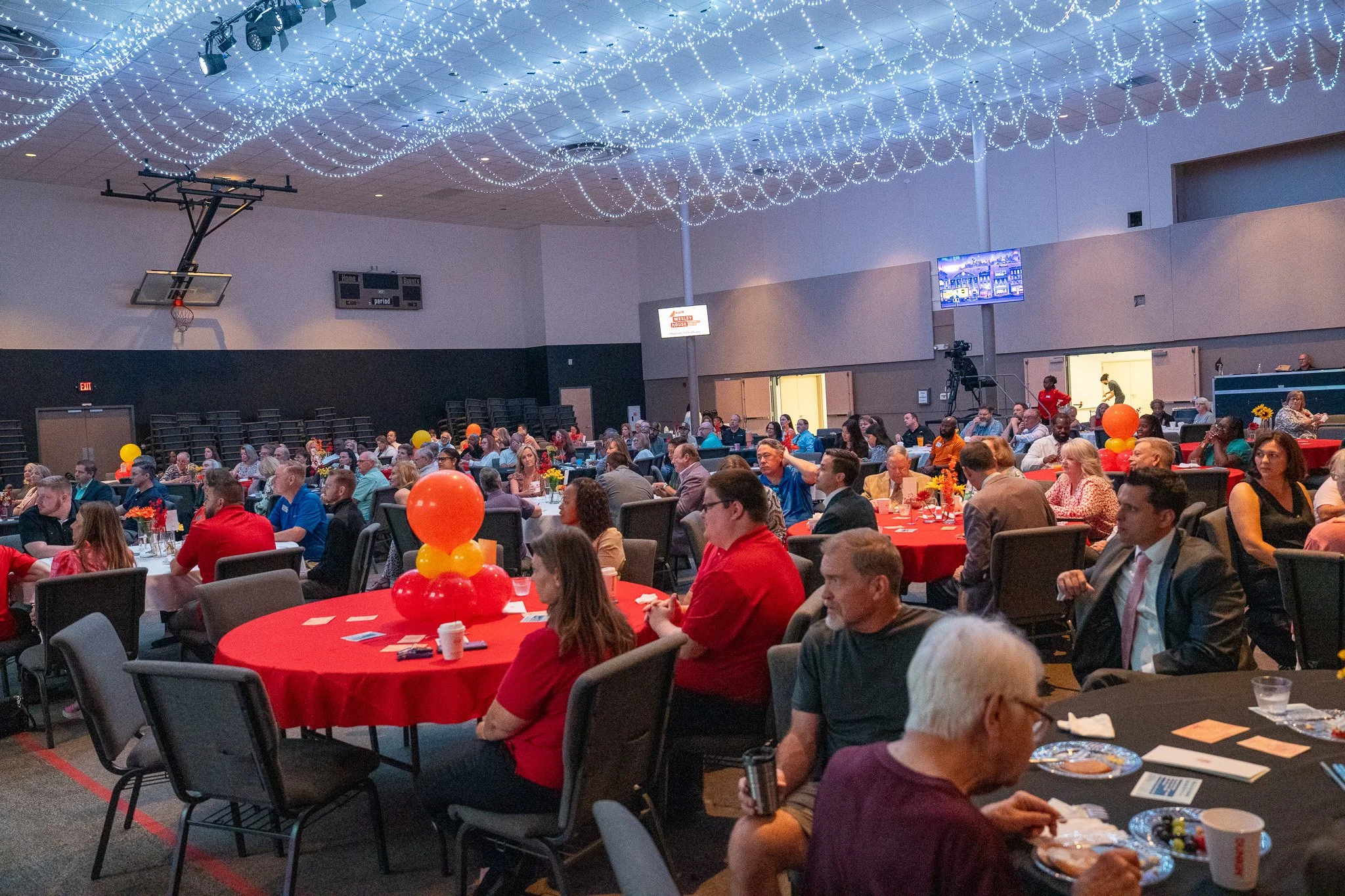 A large banquet hall with round tables decorated with red tablecloths and orange balloons, filled with attendees seated and talking. The ceiling is decorated with string lights, and there are large screens and a camera setup at the back.