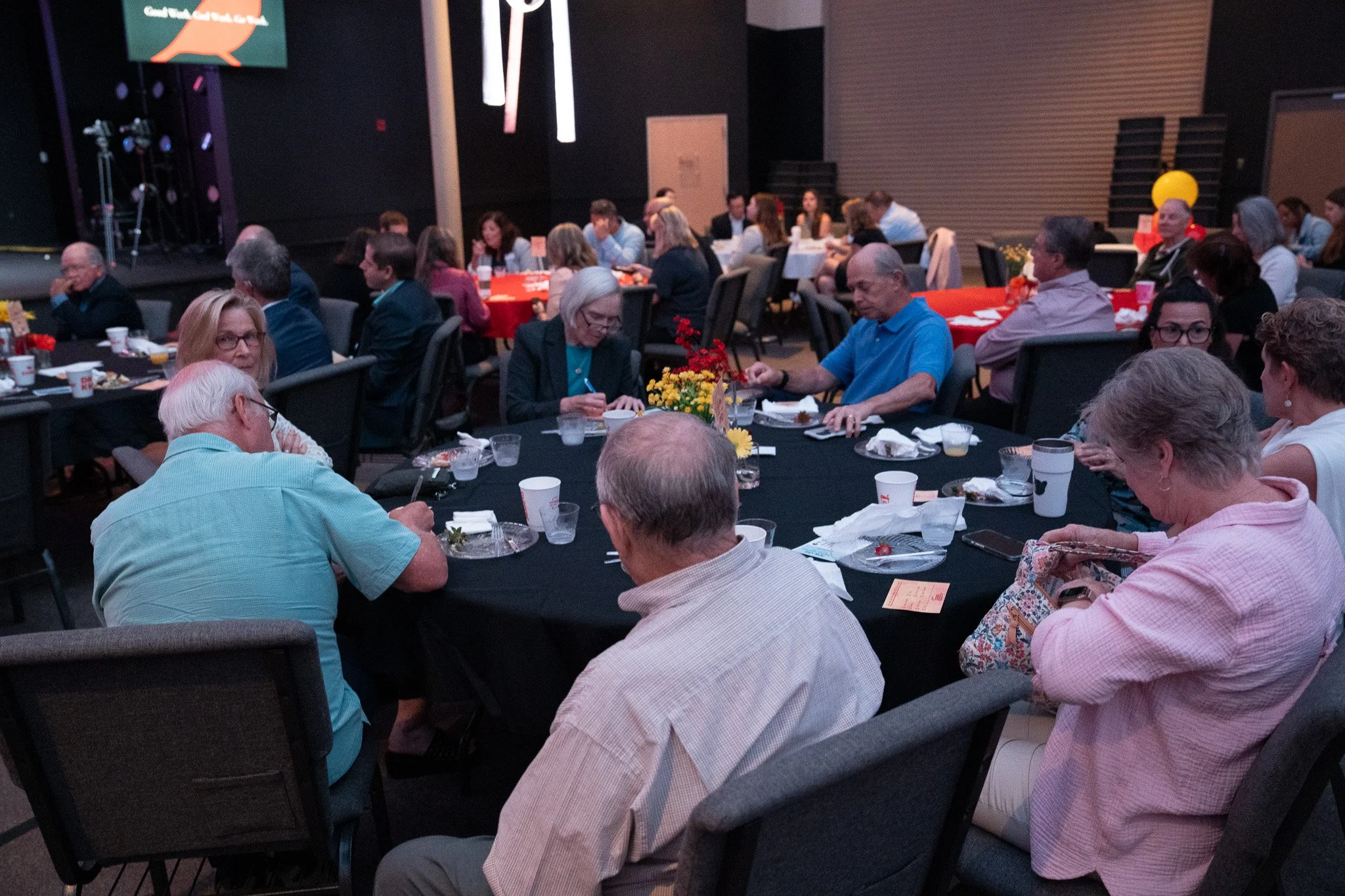 People sitting at round banquet tables with black tablecloths, eating and talking, in a dark indoor venue with a stage and a large screen in the background.