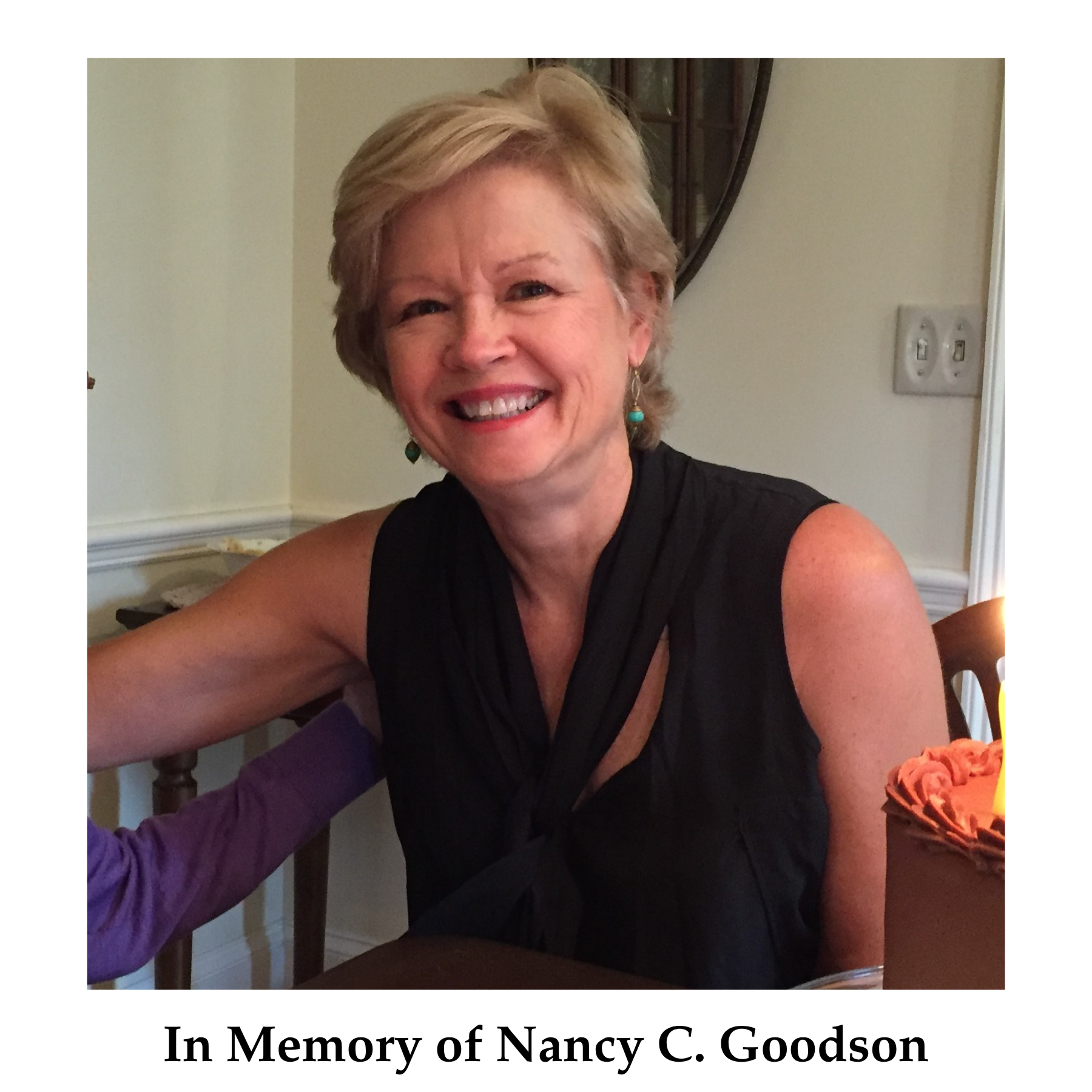 Smiling woman with short blonde hair in black sleeveless top, sitting at a table with birthday cake, celebrating Nancy C. Goodson's memory.