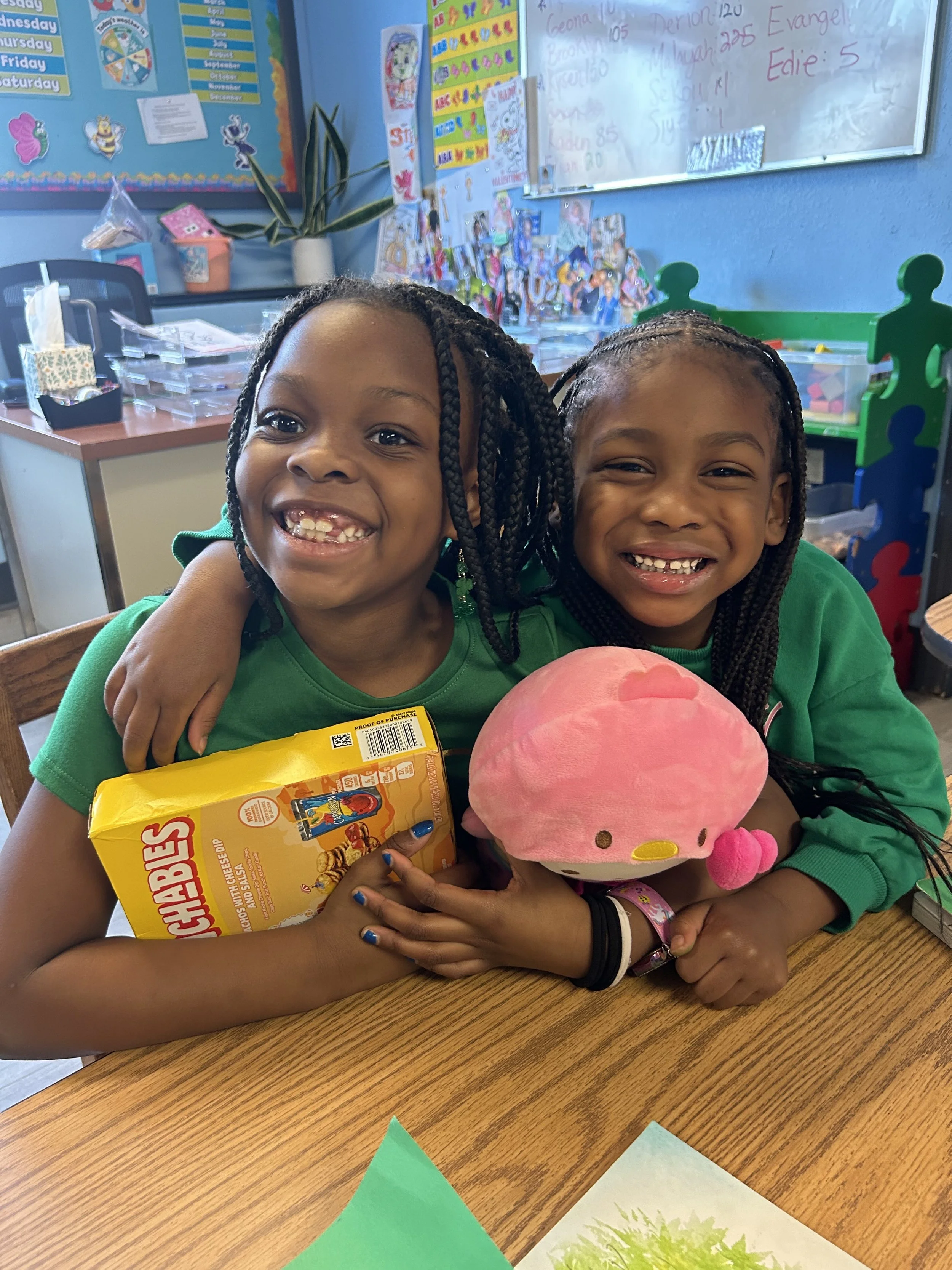 Two young girls with braided hair smiling and hugging at a classroom table, holding a pink plush toy and a box of Cheez-Its snack, with colorful classroom decorations and whiteboard in the background.